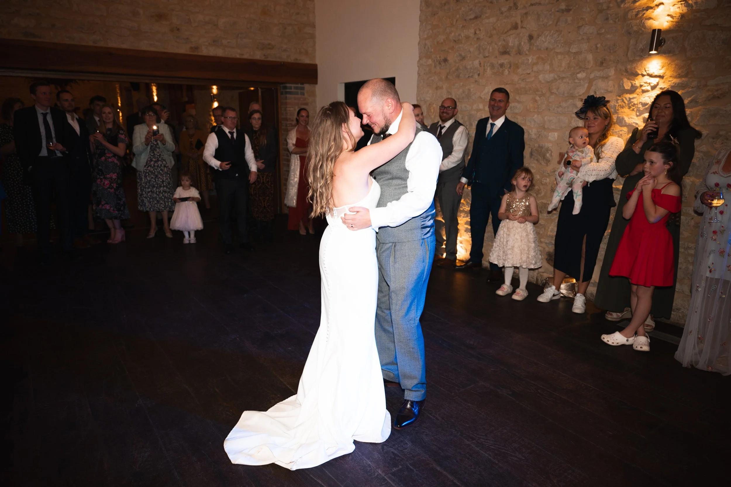 A bride and groom share their first dance at a wedding reception, surrounded by guests in a warmly lit room with brick walls.