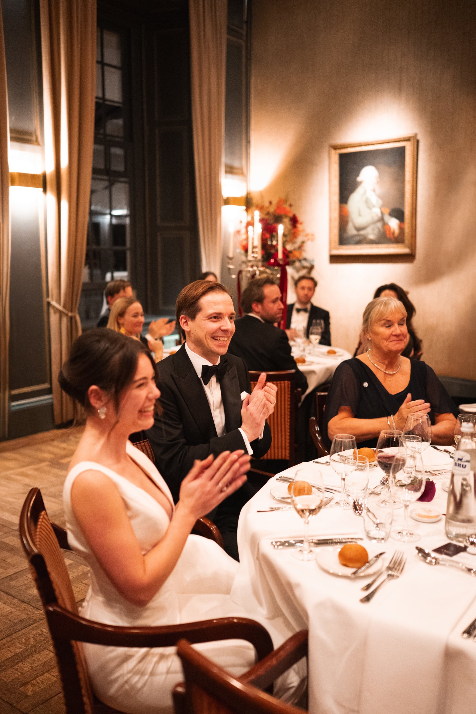 People dressed in formal attire, sitting at a round table with white tablecloth, clapping and smiling during a dinner in an elegant room with paintings and curtains.