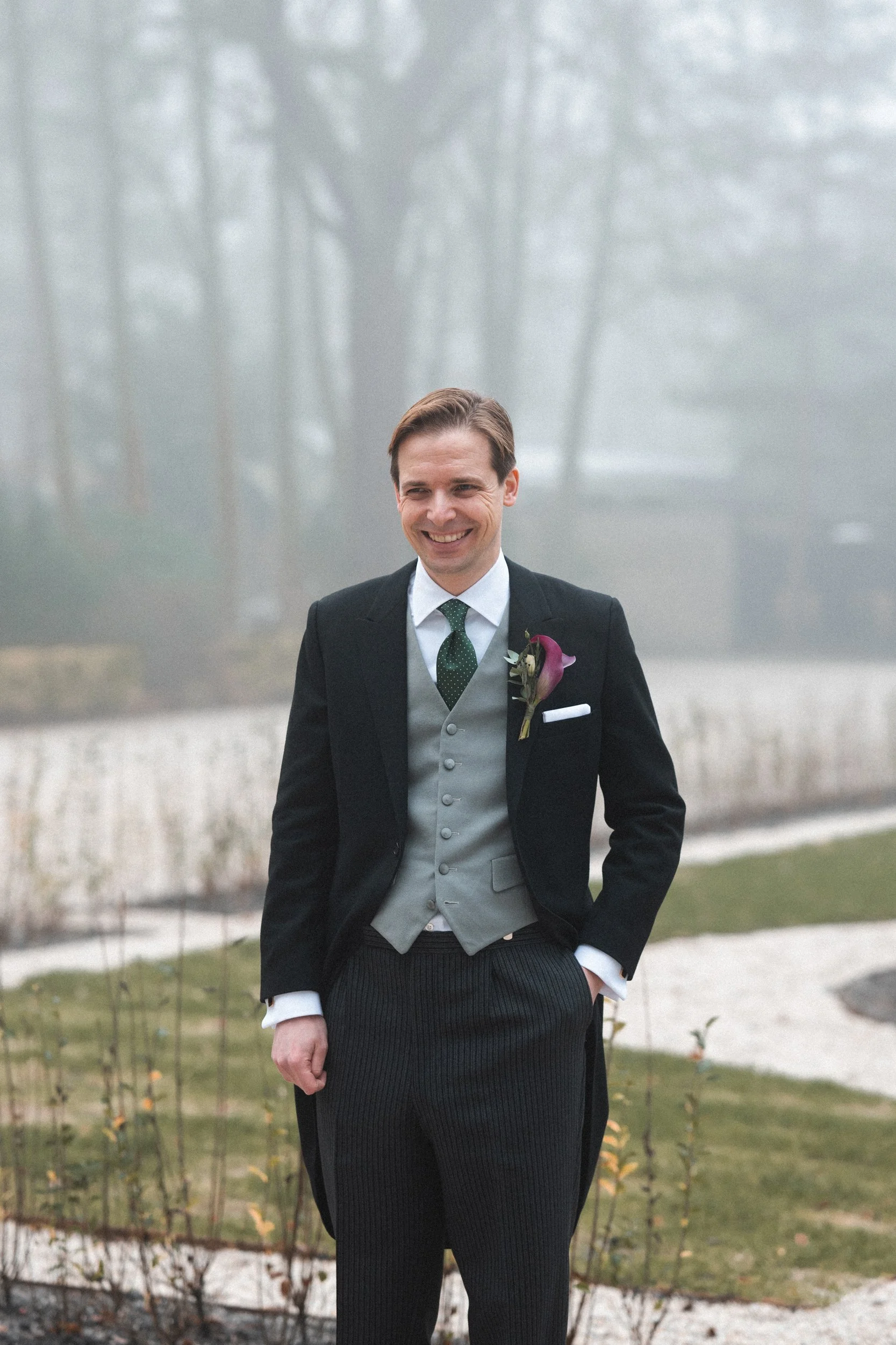 A man in a tuxedo with a boutonniere, standing outdoors in a foggy park with trees and lawns.
