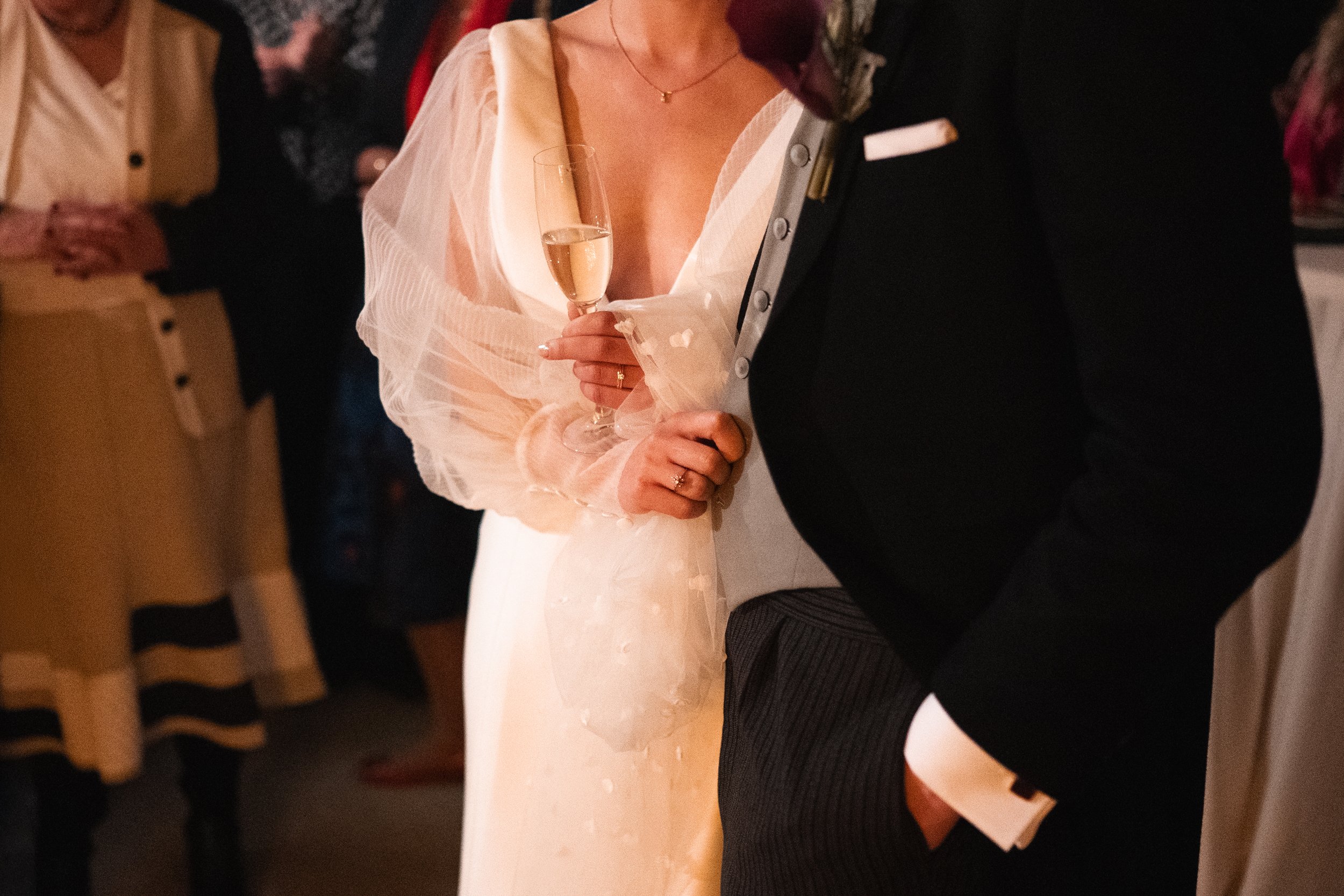 Partially visible woman in a wedding dress holding a champagne flute at a wedding reception, with other guests in the background.
