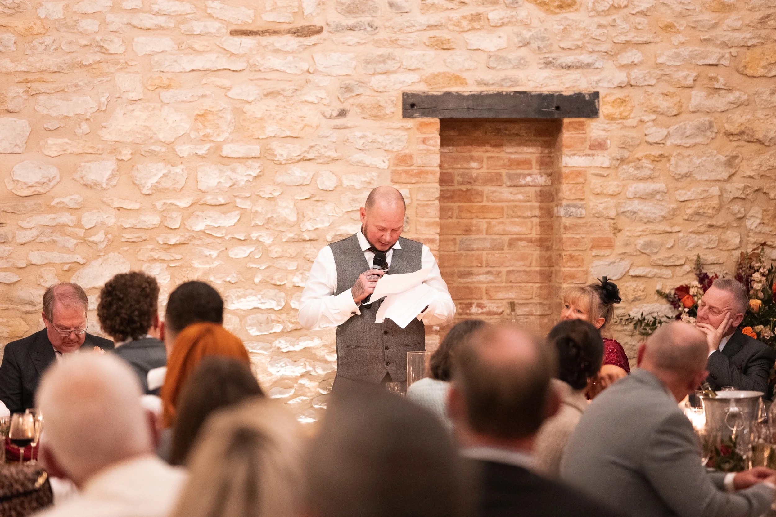 A man giving a speech at a wedding reception, standing in front of a brick wall with a black wooden beam. He is dressed in a white shirt, gray vest, and tie, holding a microphone and reading from papers. Guests seated at tables watch him, with some s