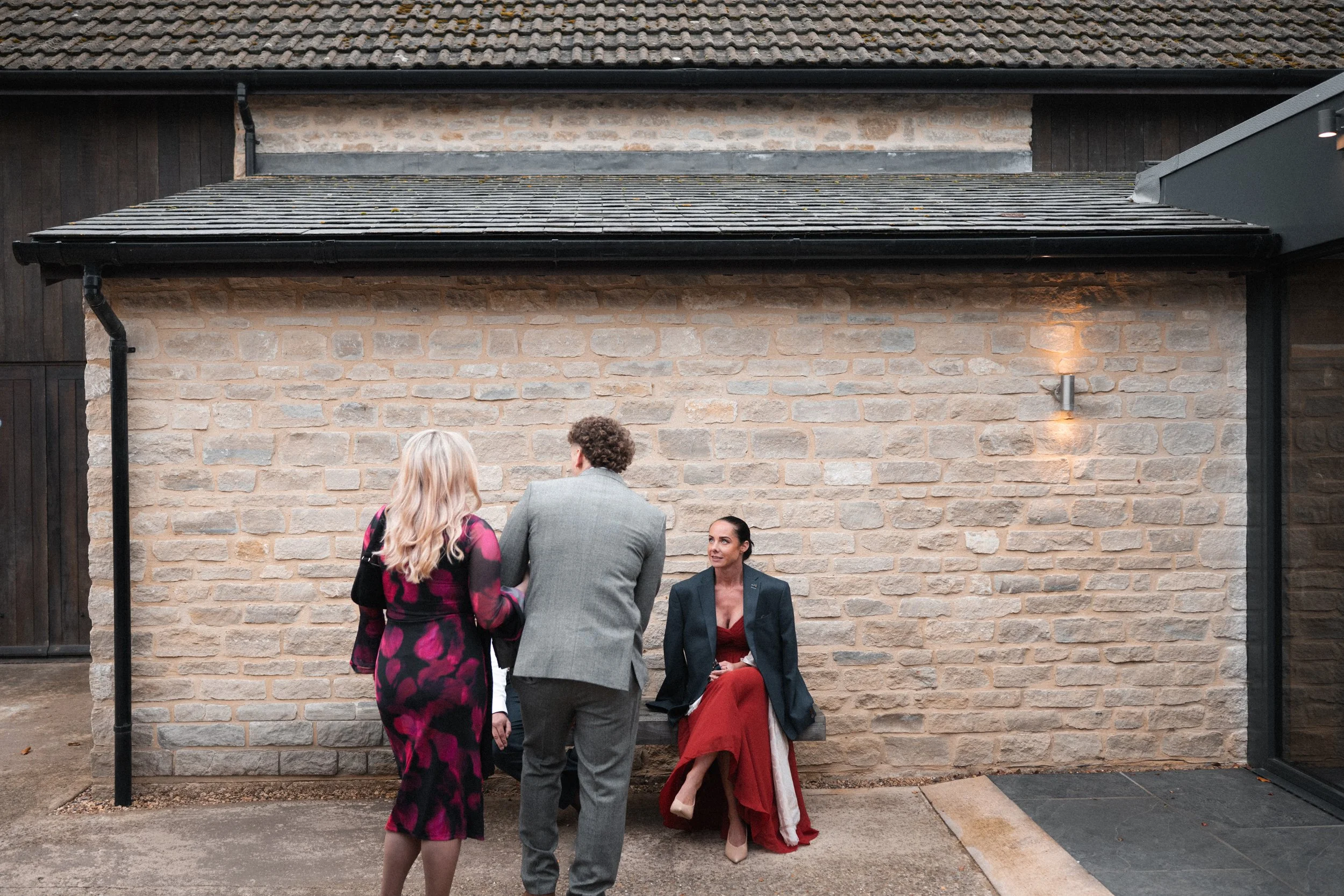 Four people engaged in conversation outside a building with a stone wall and a small bench. One woman is seated in a red dress and blazer, while the other three are standing, two women and one man.