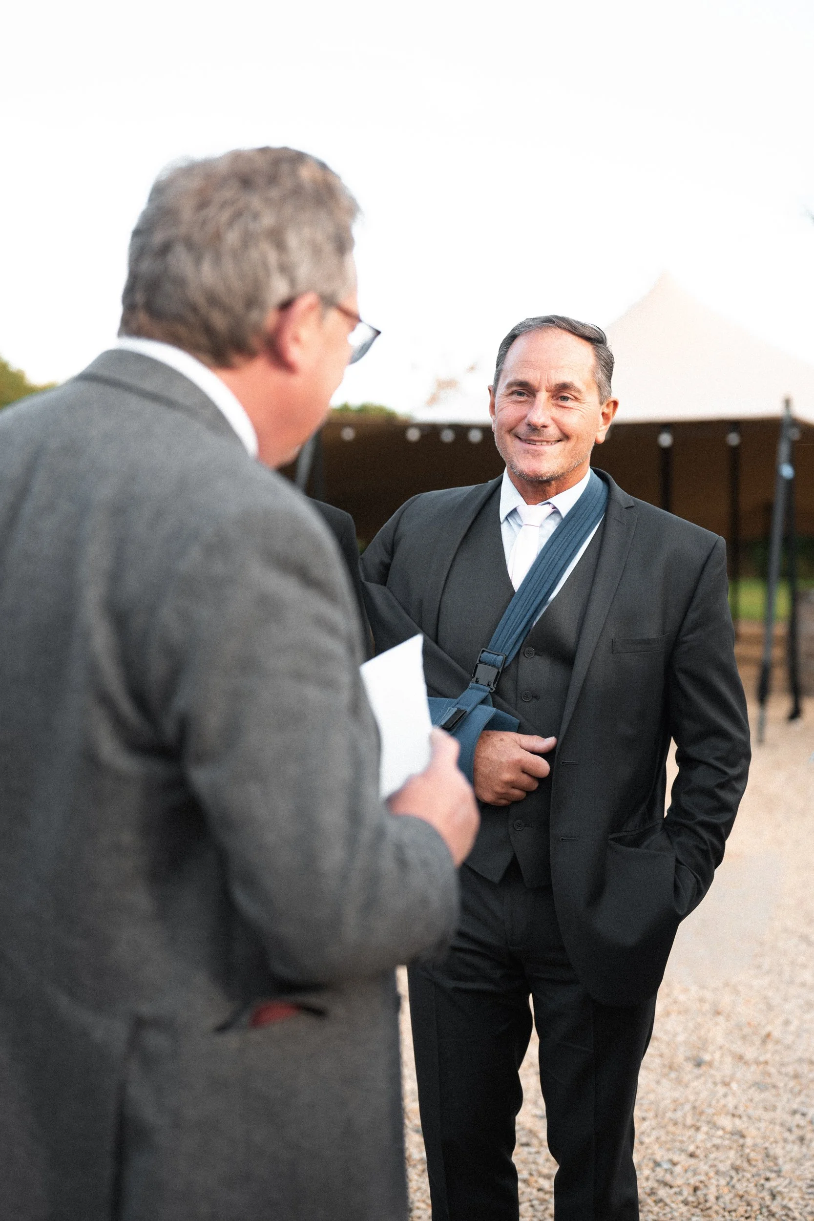Two men in suits talking outdoors with a white tent in the background.