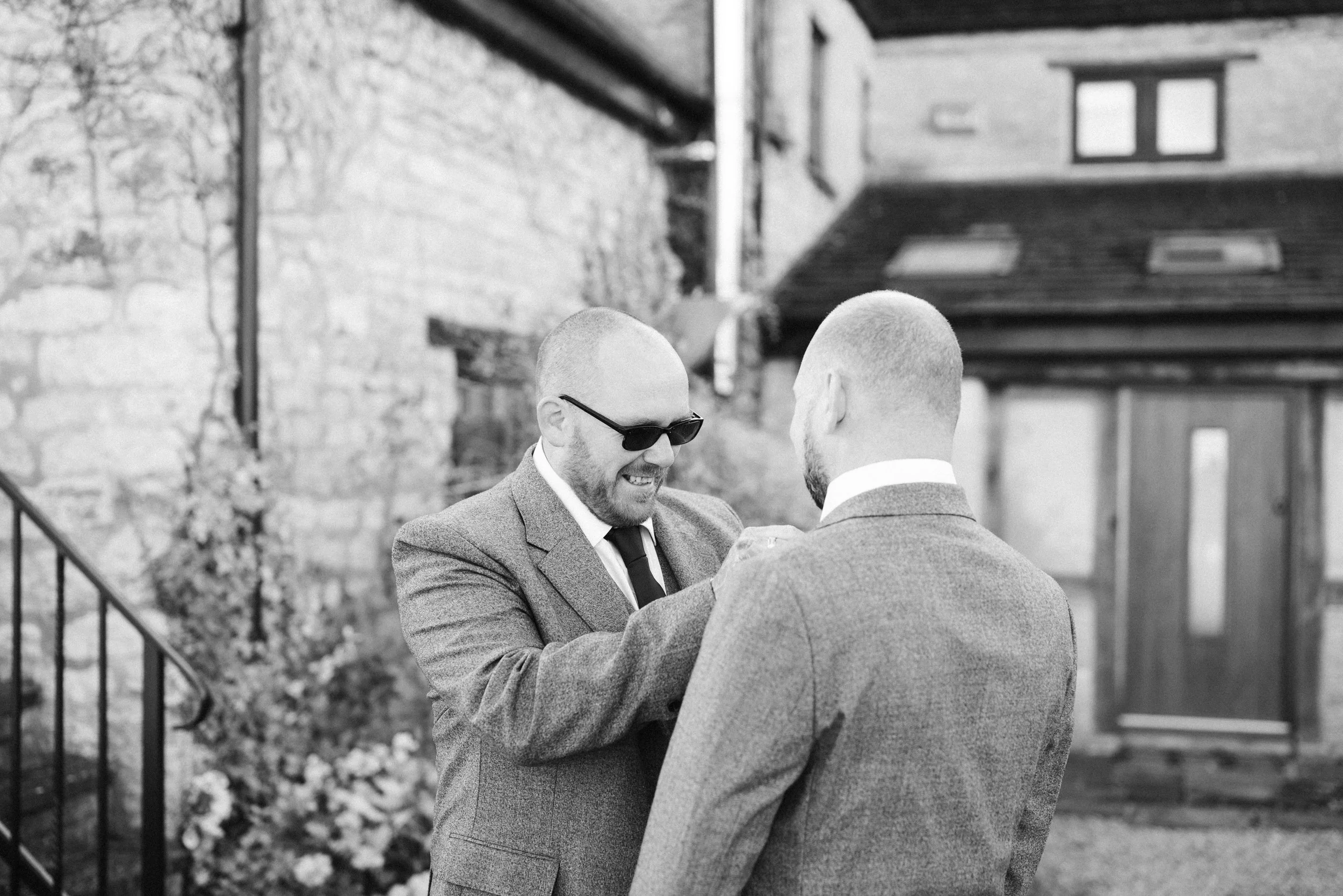 Two men in suits standing close together exchanging rings outdoors, one is smiling and wearing sunglasses.