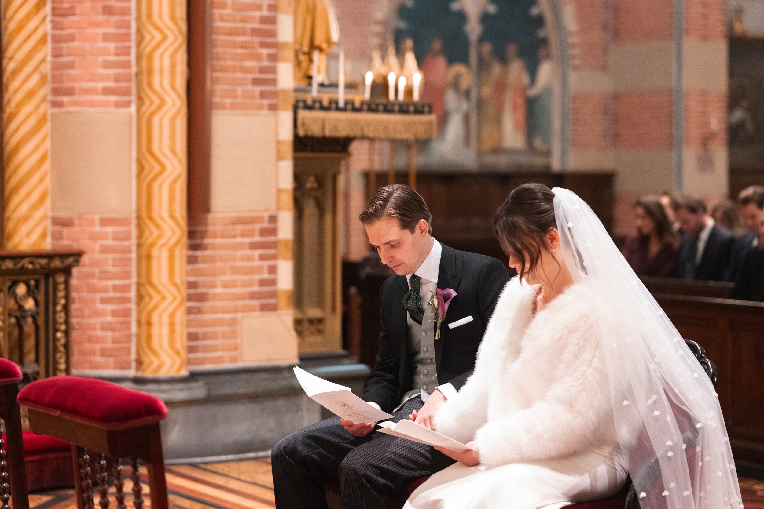 A bride and groom sitting in a church during their wedding ceremony, both with their heads bowed and reading from papers, with candles and religious paintings in the background.