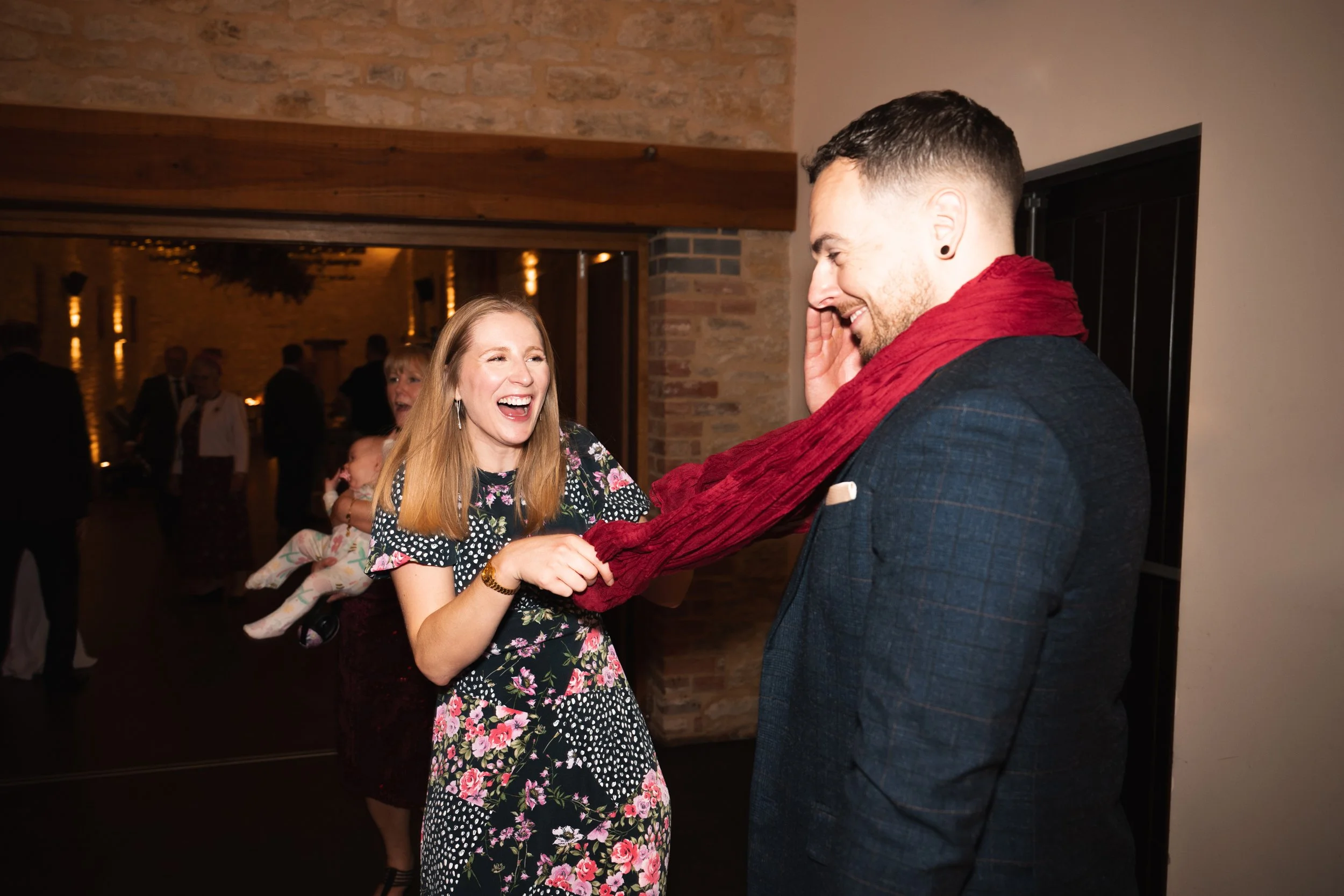 A woman with red hair wearing a floral dress is smiling and laughing while playfully tugging a red scarf wrapped around a man's neck. The man, with short dark hair and a beard, is dressed in a dark plaid suit and is smiling with his hand near his che