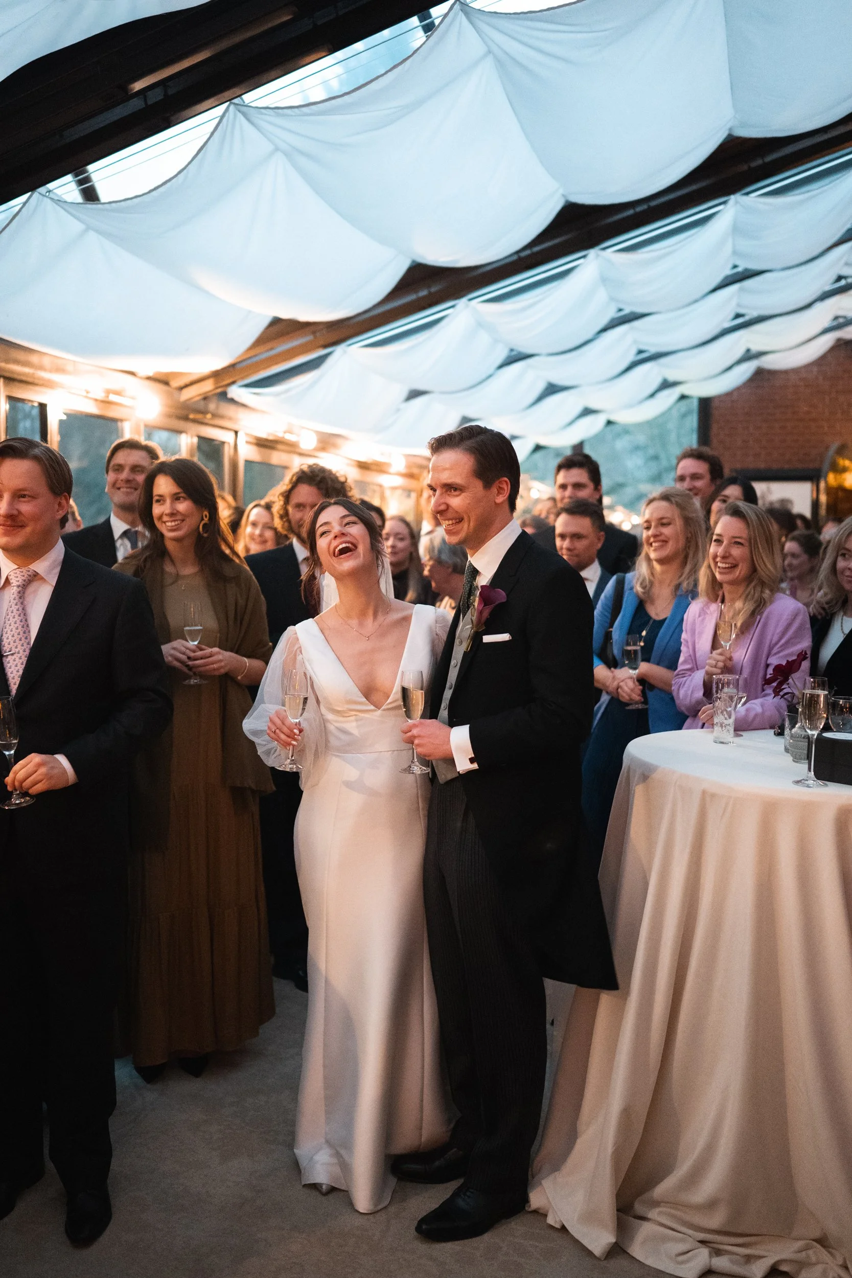 Wedding reception with a bride and groom smiling and holding champagne glasses, surrounded by friends in elegant attire under decorated ceiling panels.