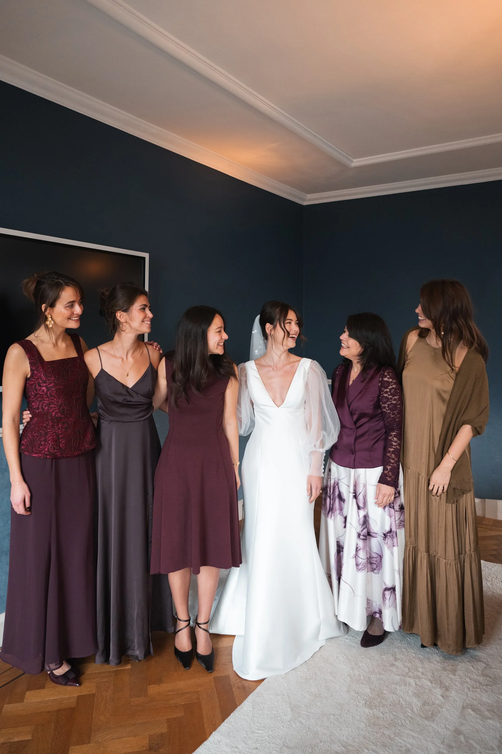 A bride with dark hair in a white wedding dress standing with five women in formal dresses, smiling and looking at each other inside a room with a dark blue wall, wooden floor, and a large screen on the wall.