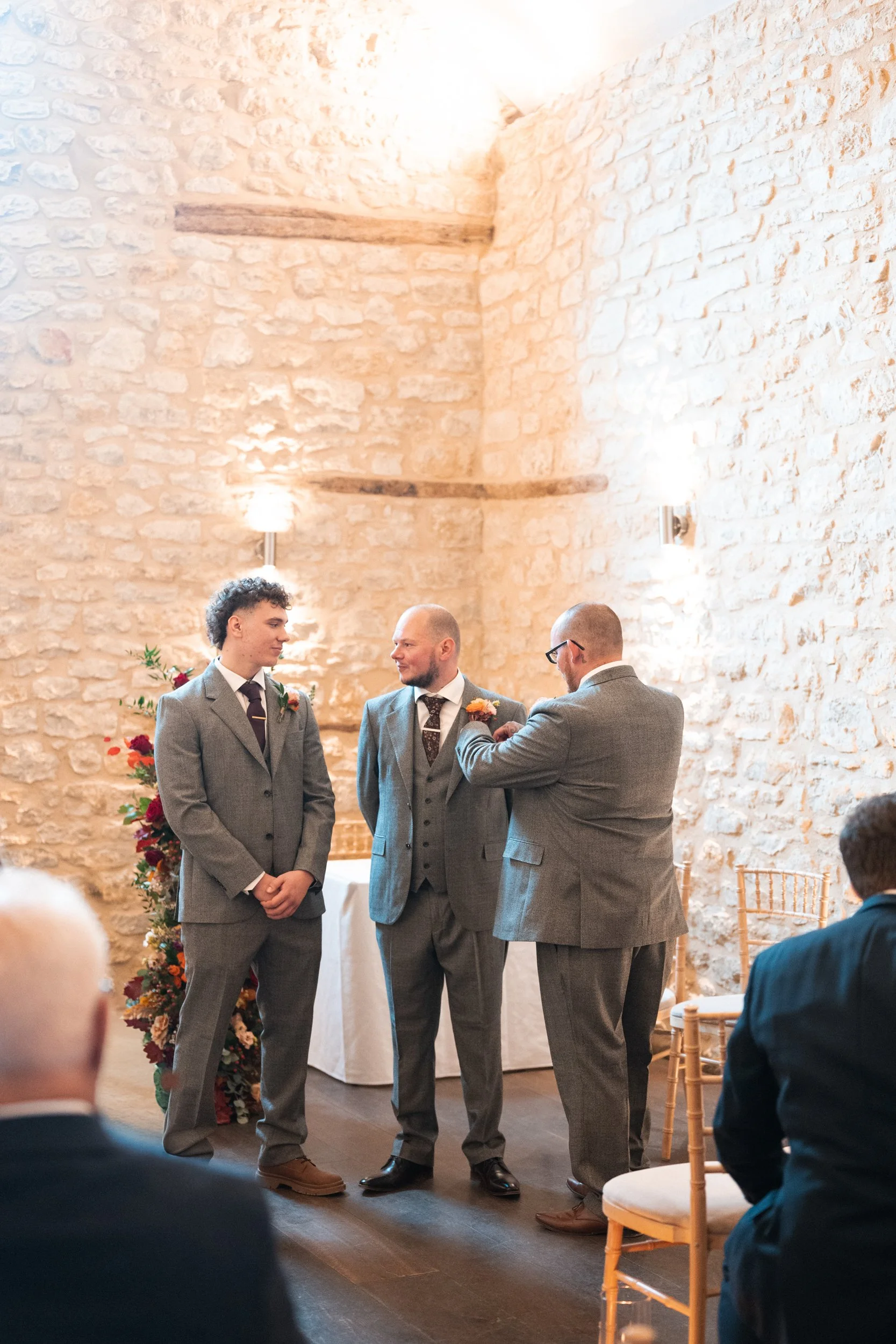 A wedding ceremony with two grooms and an officiant in a rustic stone-walled room decorated with flowers.