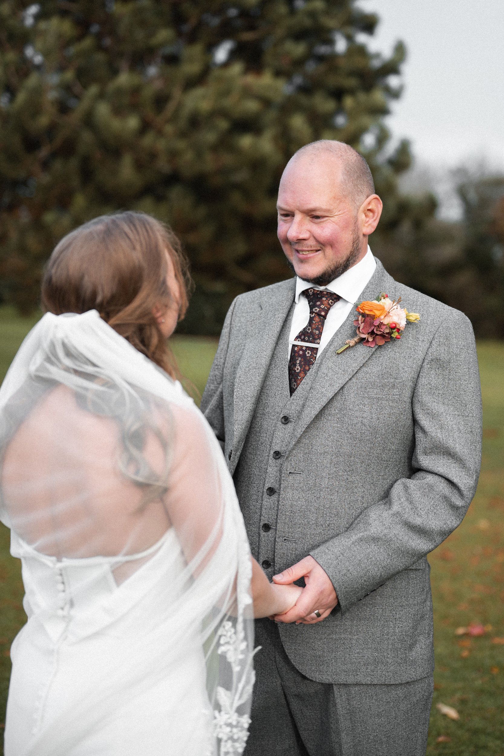 A couple getting married outdoors, holding hands, with the groom smiling and the bride with her back turned, wearing a white wedding dress and veil, and a man in a gray suit with a boutonniere standing in front of her.