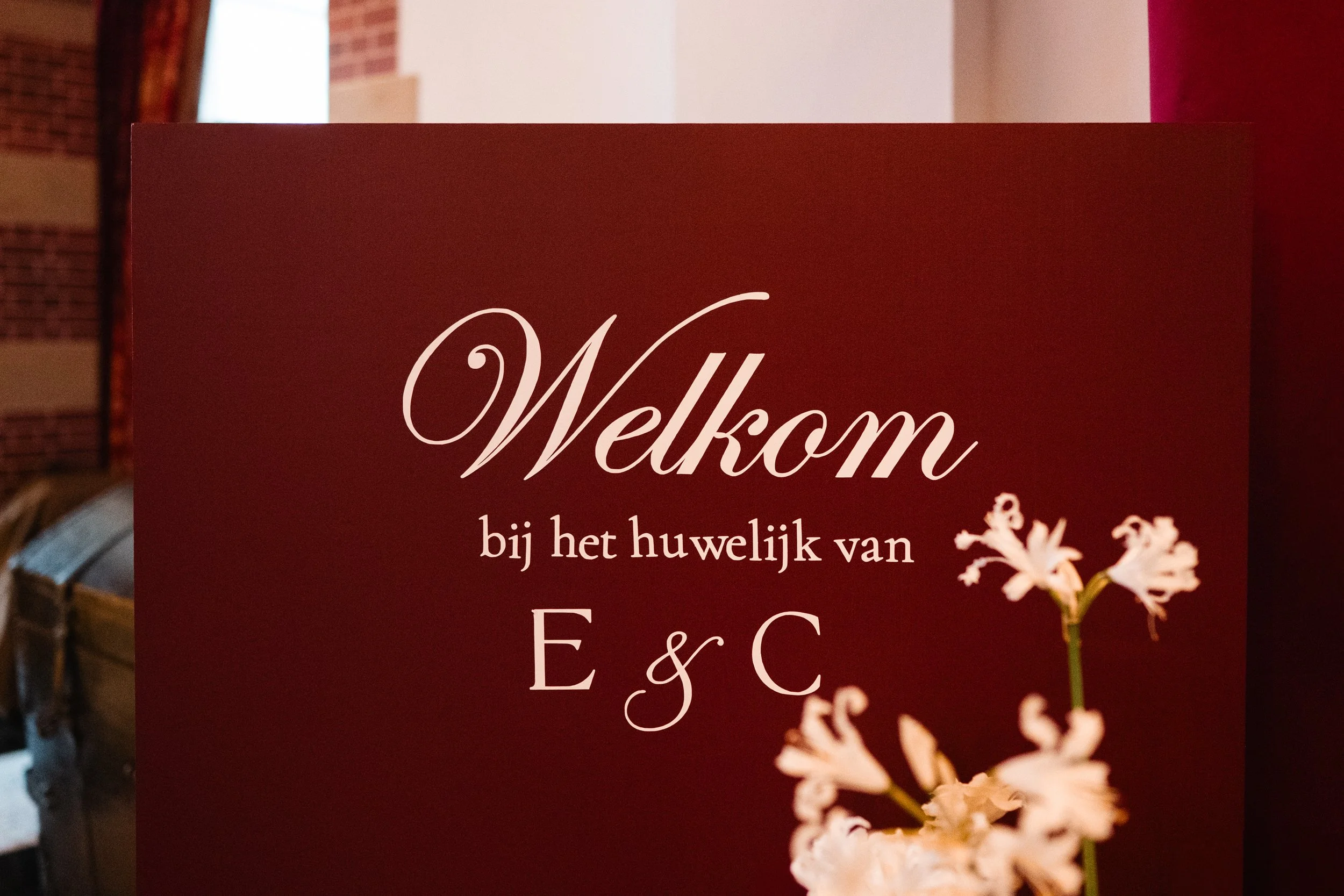 A wedding welcome sign in Dutch that says 'Welkom bij het huwelijk van E & C' with pink flowers on a maroon background