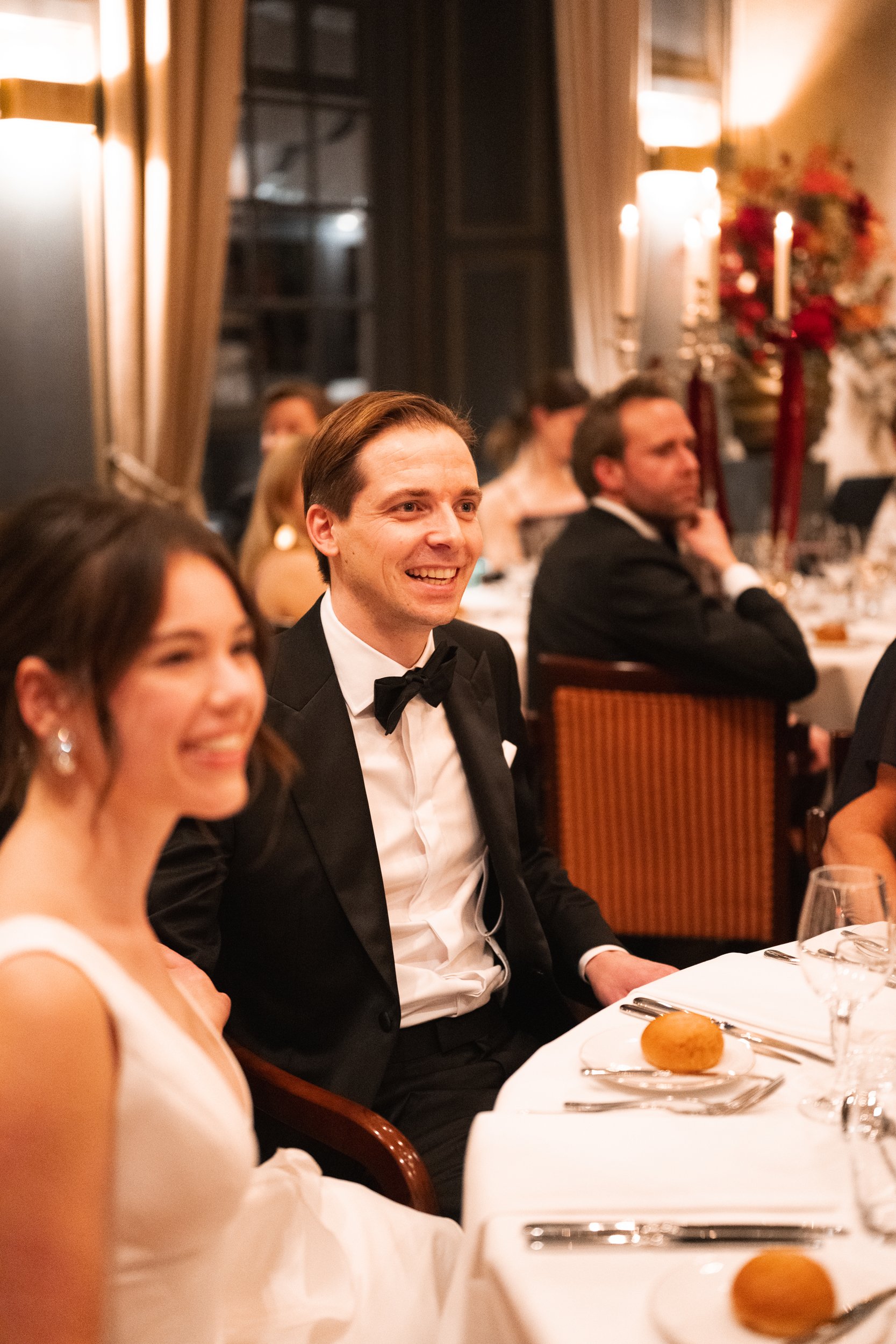 A man in a tuxedo with a bow tie and a woman in a white dress sitting at a formal dinner table, smiling, in an elegant room with warm lighting and floral decorations.