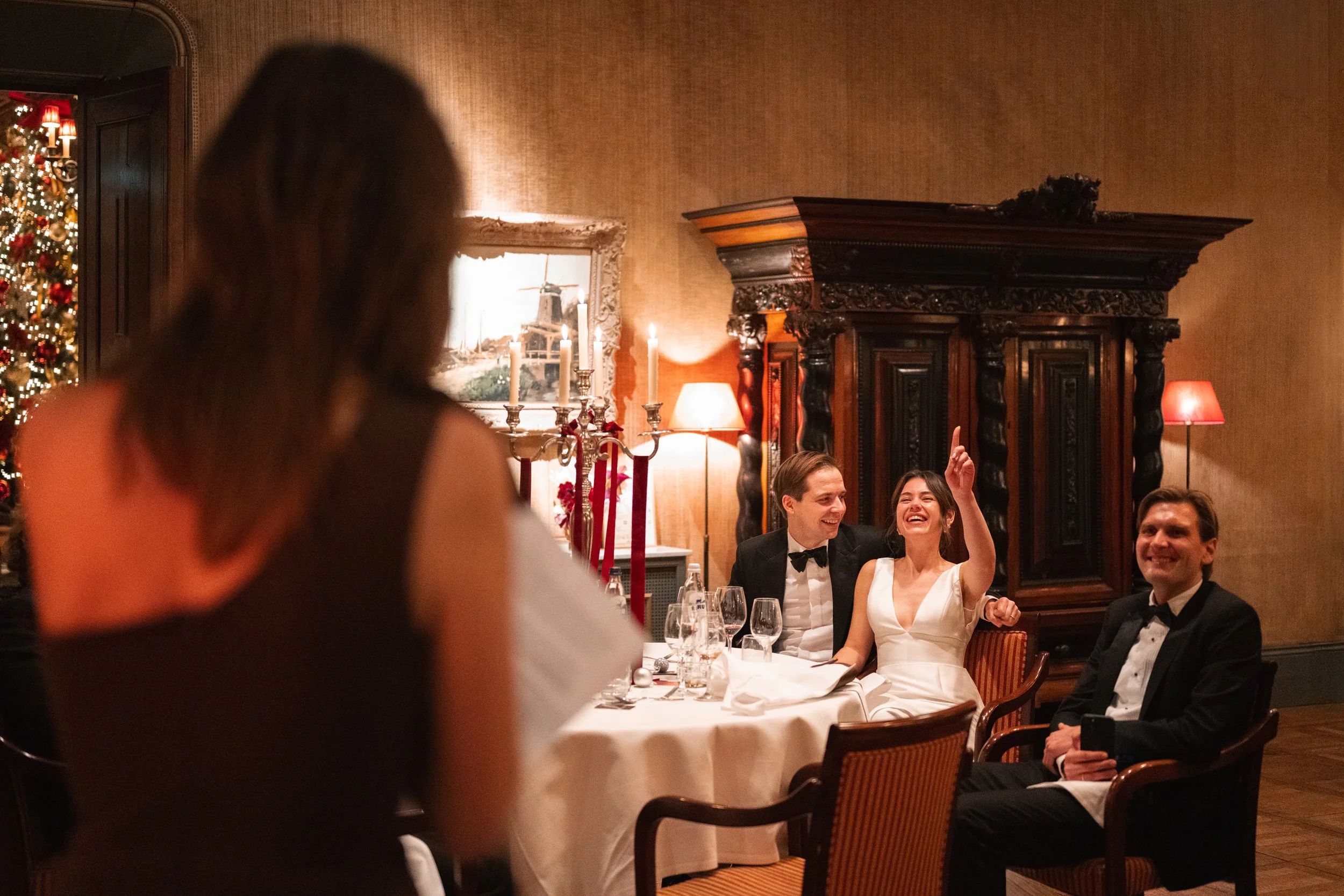 A woman in a black dress is standing in front of a group of people at a festive dinner table. Four people are seated, smiling and one woman is laughing or talking with her hand raised. The setting is decorated with Christmas ornaments, candles, and w
