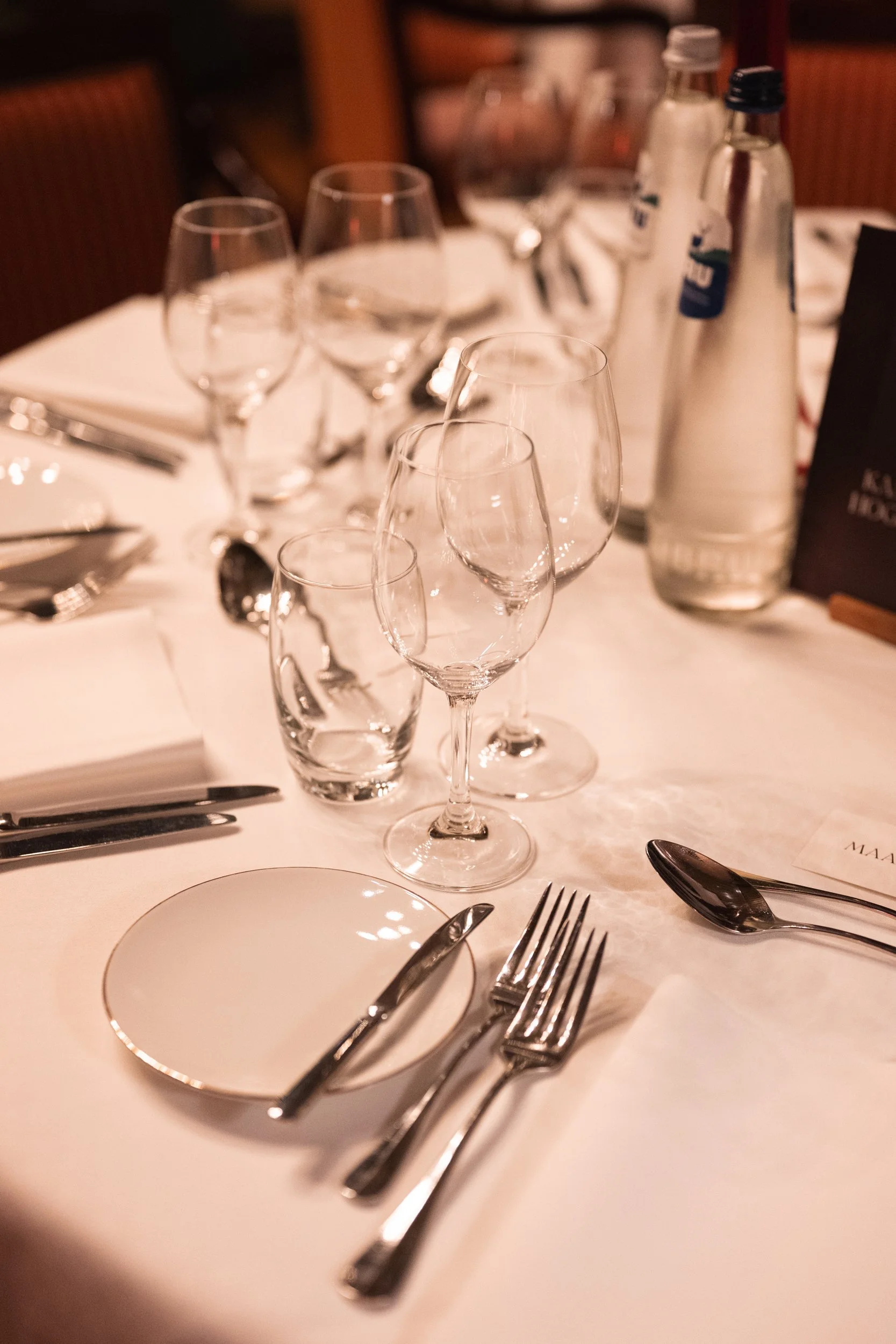 Elegant table setting with multiple wine glasses, water glasses, silverware, a small plate, and bottled water on a white tablecloth in a restaurant.