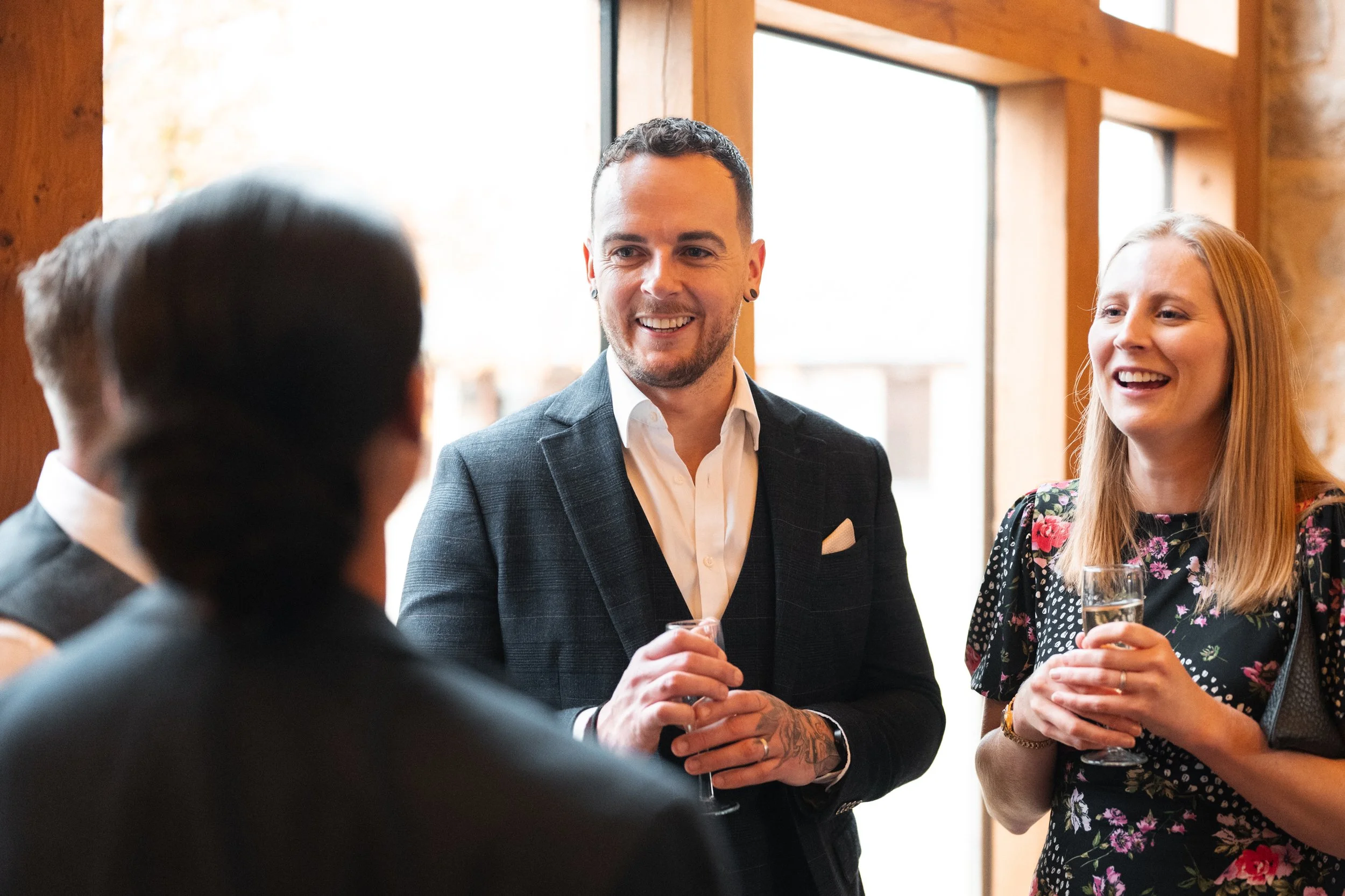 Groups of people socializing at an indoor event, with a man and woman smiling and holding glasses of champagne.