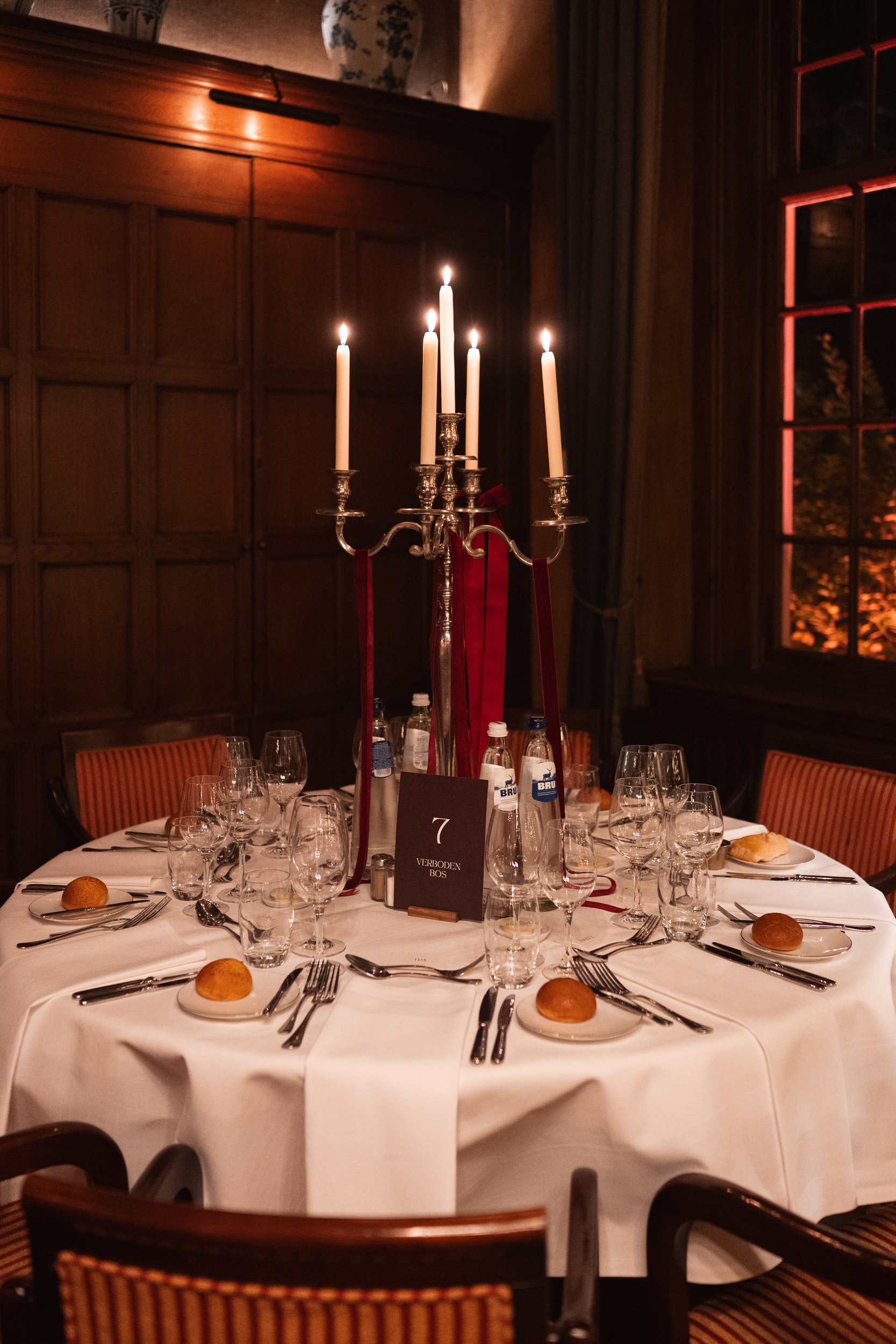Elegant round dining table set for a formal dinner with white tablecloth, multiple wine glasses, water bottles, bread rolls, silverware, and a black table number sign, illuminated by a tall candelabrum with lit candles in a dimly lit wood-paneled roo