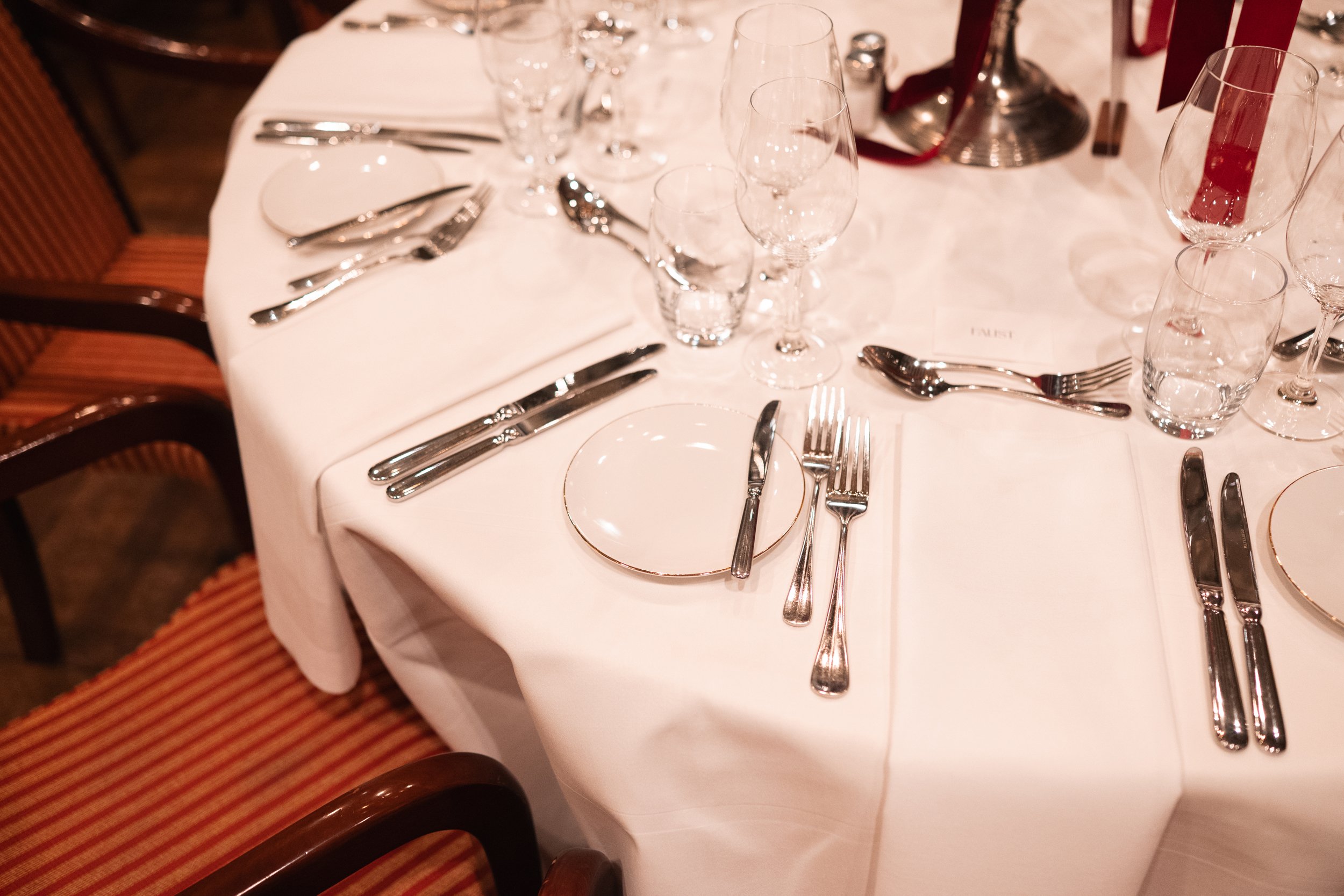 Round dining table set with white tablecloth, multiple glasses including wine and water glasses, white plates with gold rim, silverware arranged for a formal dinner, and chairs with red and gold striped upholstery.