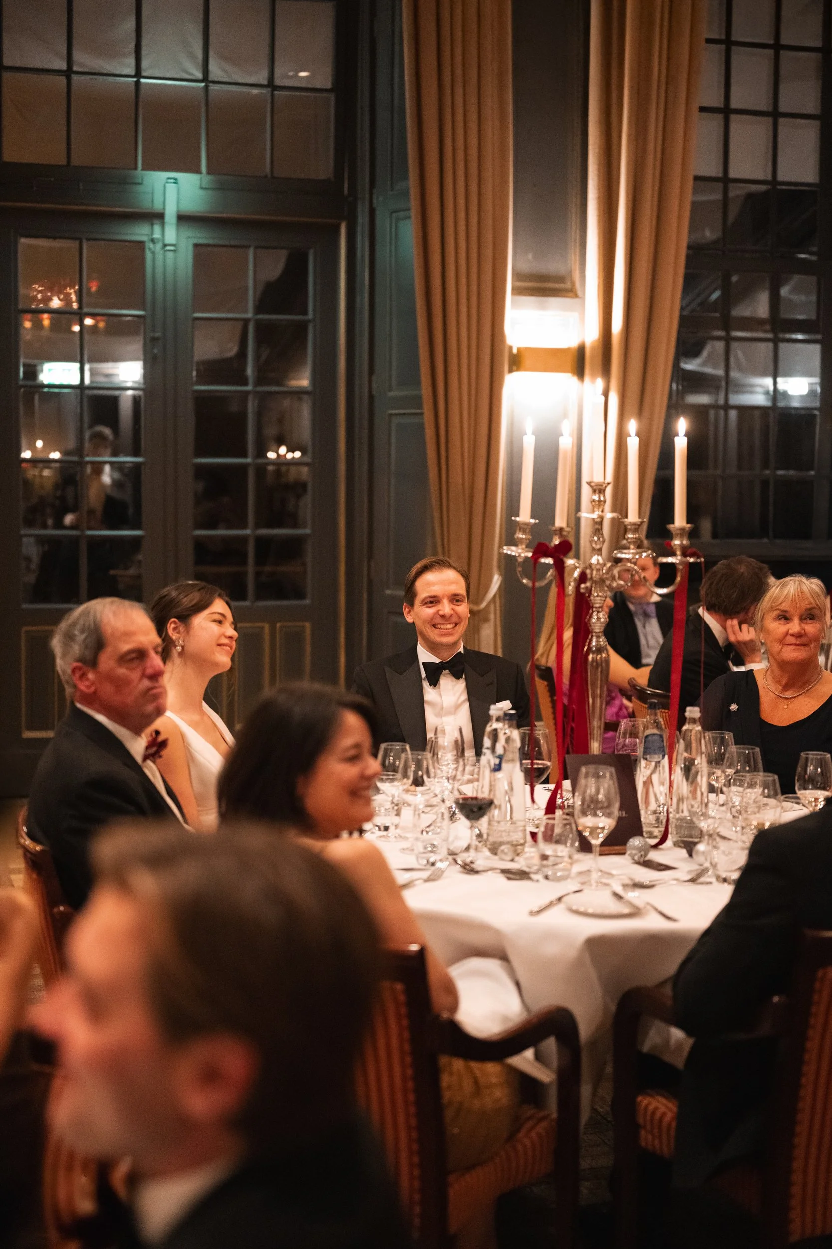 People sitting at a dining table with elegant wine glasses and a large candelabra with lit candles, in a sophisticated, warmly lit event space with tall windows and curtains.