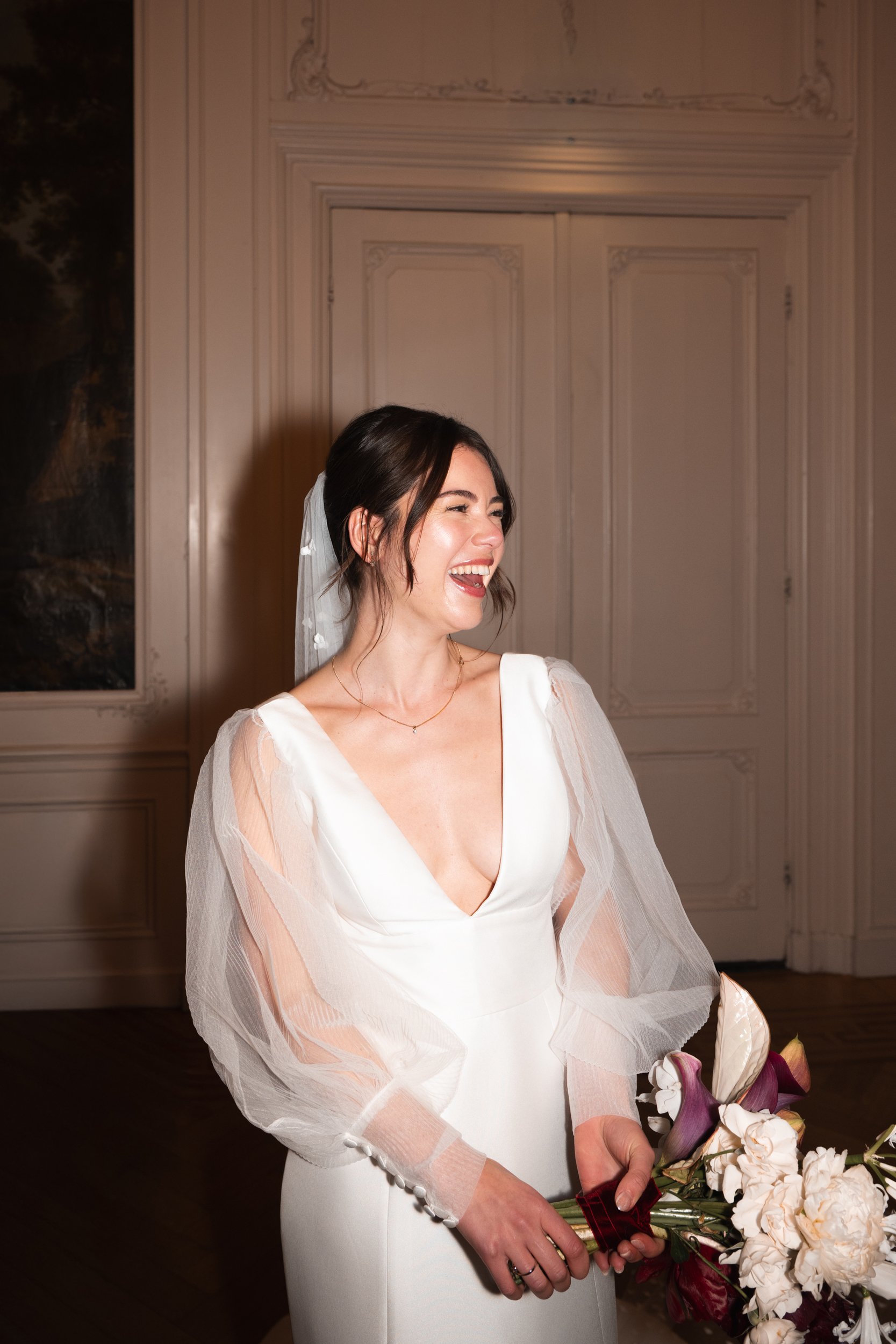 A woman in a white wedding dress with sheer puffed sleeves, holding a bouquet of flowers, smiling and laughing indoors.