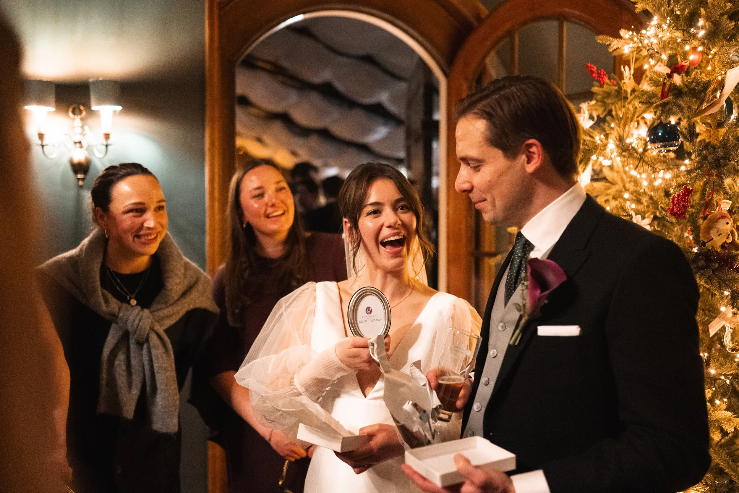 A group of people celebrating Christmas indoors, with a decorated Christmas tree and festive lighting in the background. A woman in a white dress is laughing while talking to a man in a black suit who is holding a gift box. Two other women are standi