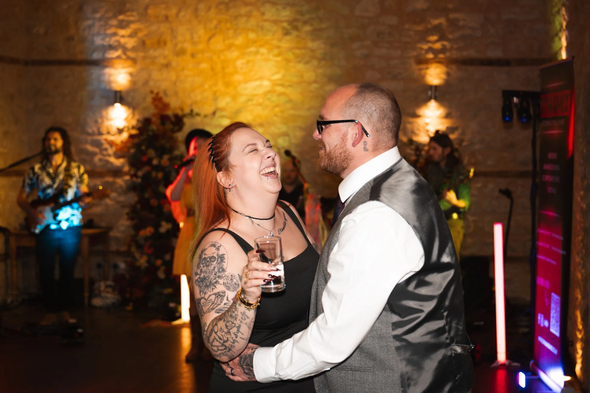 A happy woman with tattoos, holding a glass, and a man in semi-formal attire, sharing a joyful moment while dancing at an indoor party or celebration with live music in the background.