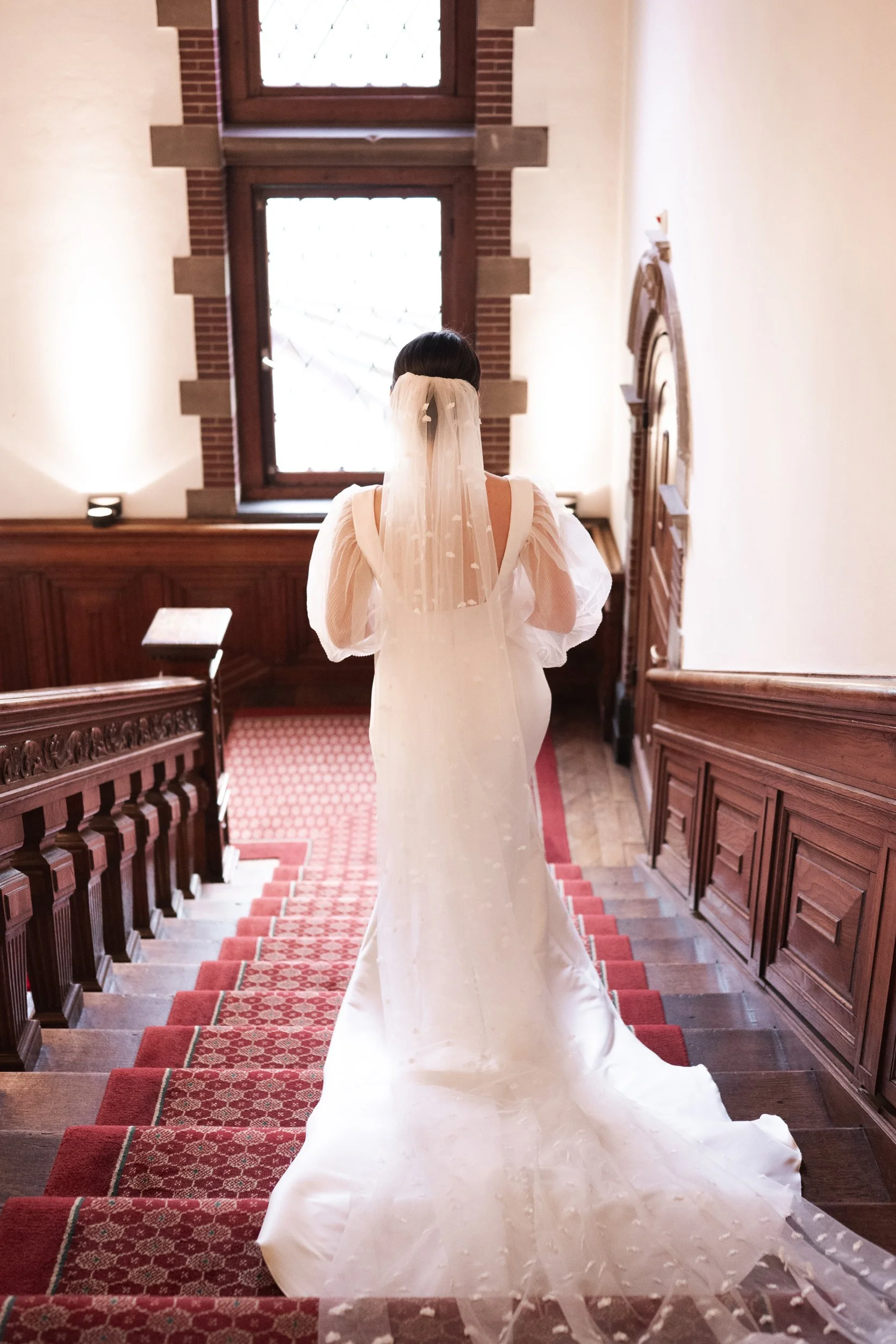 A woman in a white wedding dress with puffy sleeves and a long train standing at the top of a wooden staircase, facing a window with brick and wood framing.