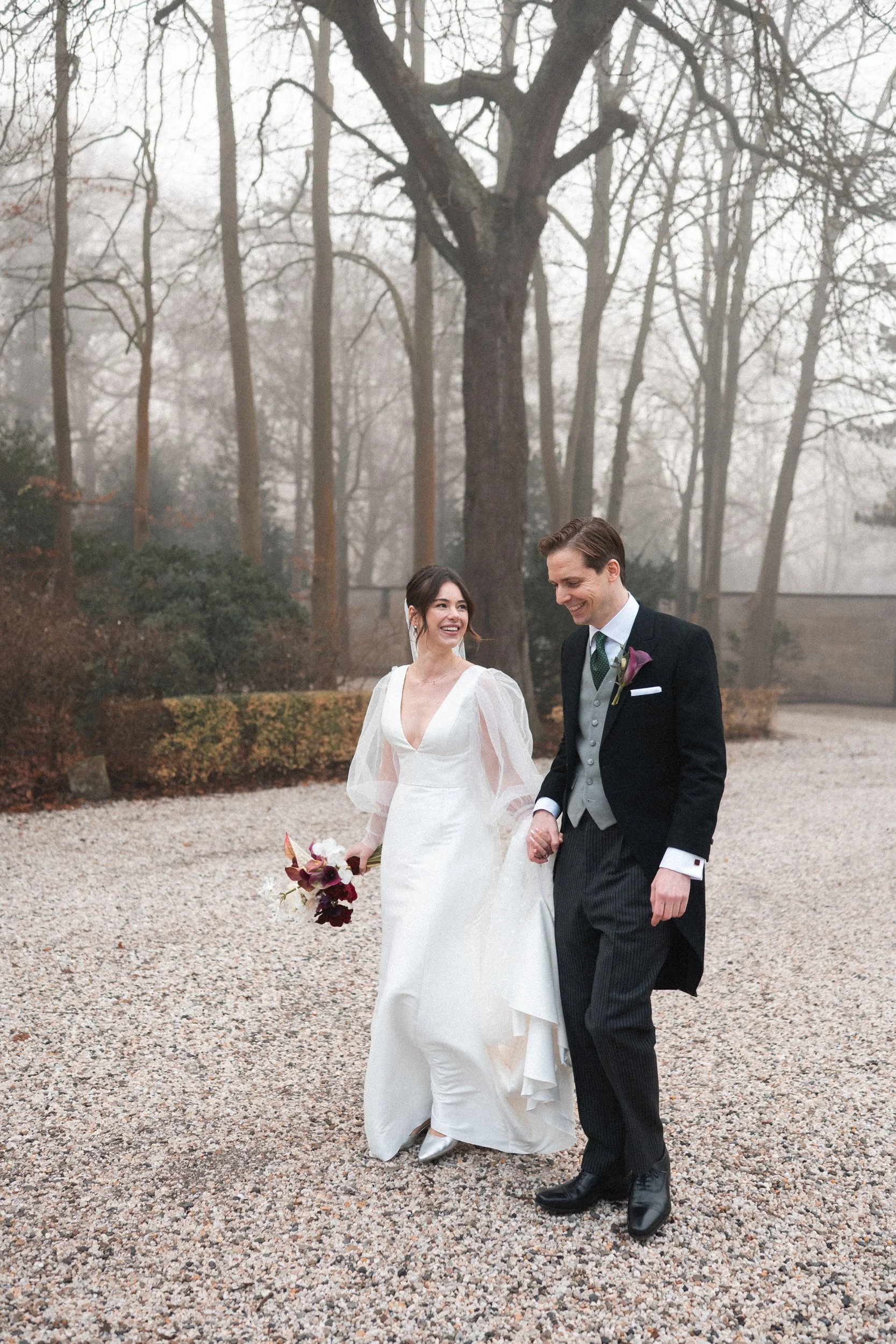 A newlywed couple walking hand in hand outdoors on a gravel path with trees in the background, smiling and enjoying their wedding day.