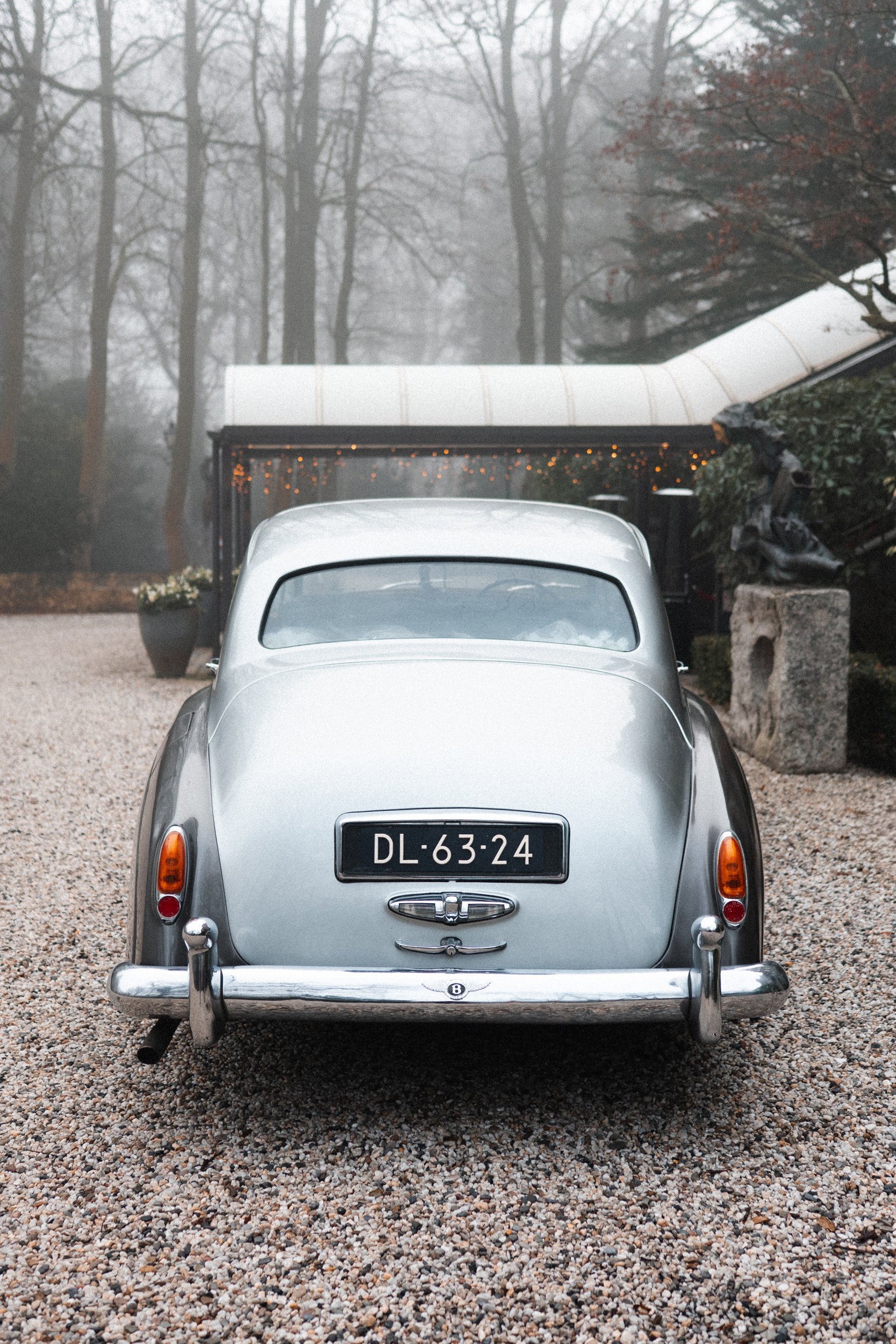 A vintage silver car is parked on a gravel driveway in front of a building with a covered porch. The background features a foggy, leafless forest, and the car's license plate reads 'DL-63-24'.
