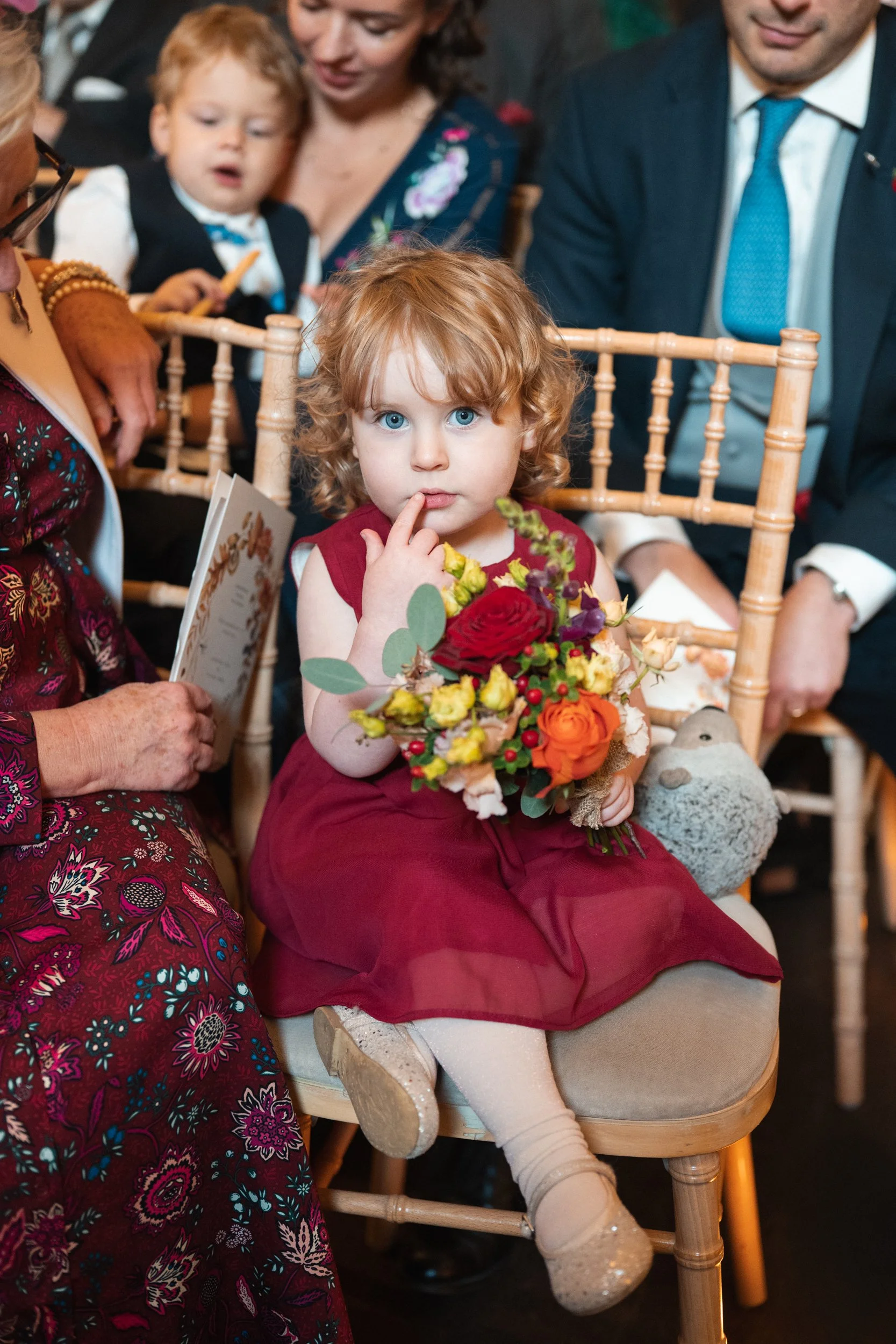 A young girl with curly red hair and blue eyes sitting on a chair, holding a colorful bouquet of flowers, with her finger near her lips at a formal event.