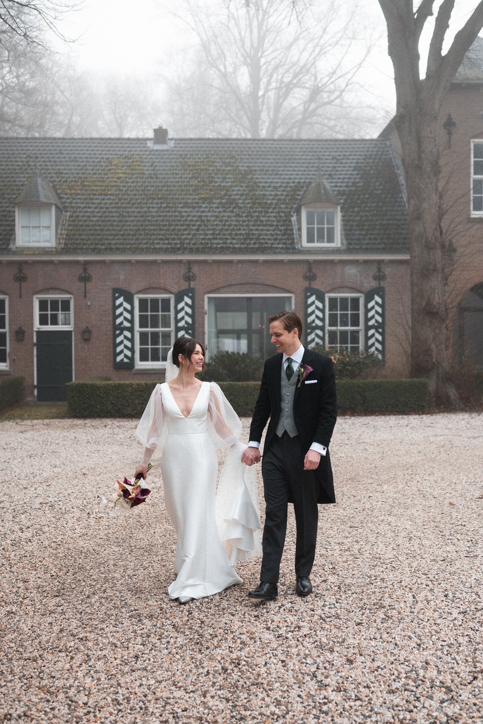 A bride and groom holding hands and walking outdoors in front of a rustic brick building with black and white striped shutters, during a wedding celebration.