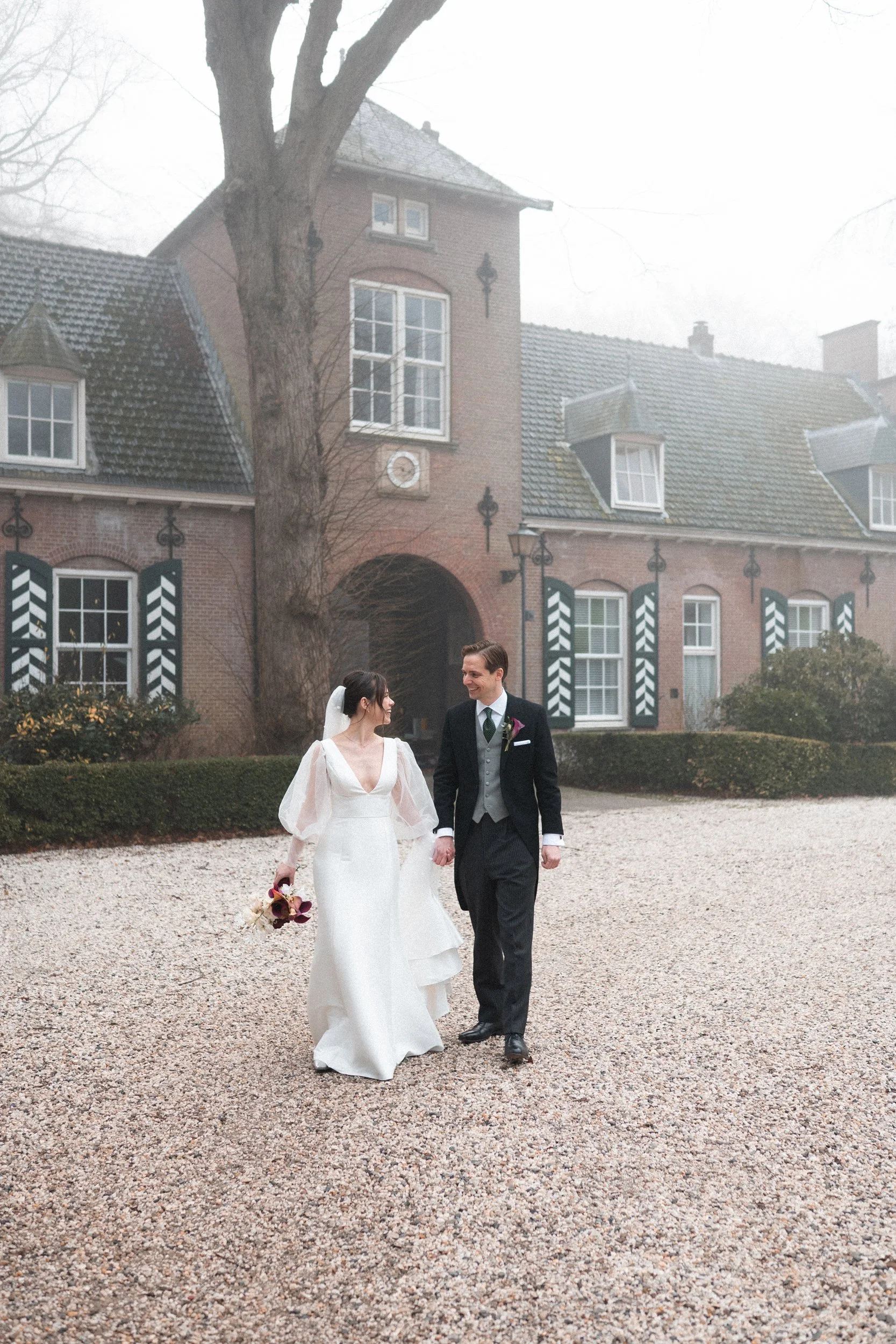 A bride and groom walking hand in hand outside on a gravel pathway in front of a large brick house with black and white shutters. The bride is holding a bouquet and wearing a white wedding dress, while the groom is in a black suit with a gray vest an