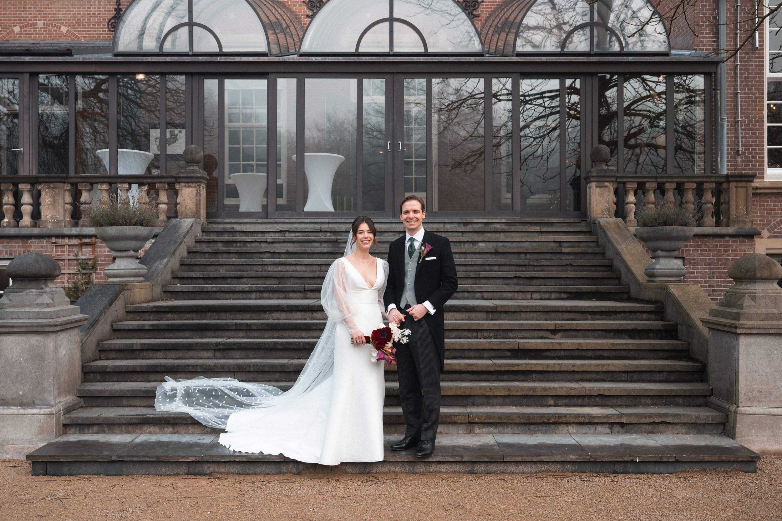 A bride and groom standing on outdoor stairs in front of a glass building, smiling and holding hands, with the bride in a white wedding dress and the groom in a black suit.