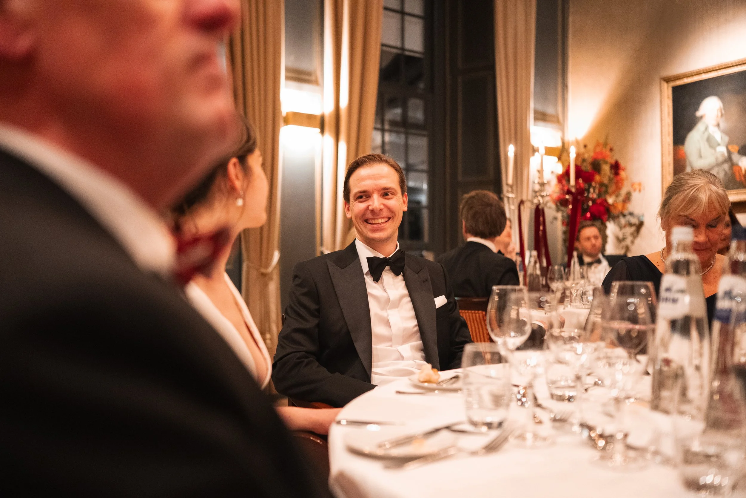 A man in a tuxedo with a bow tie smiling at a formal dinner event in an elegant decorated room.