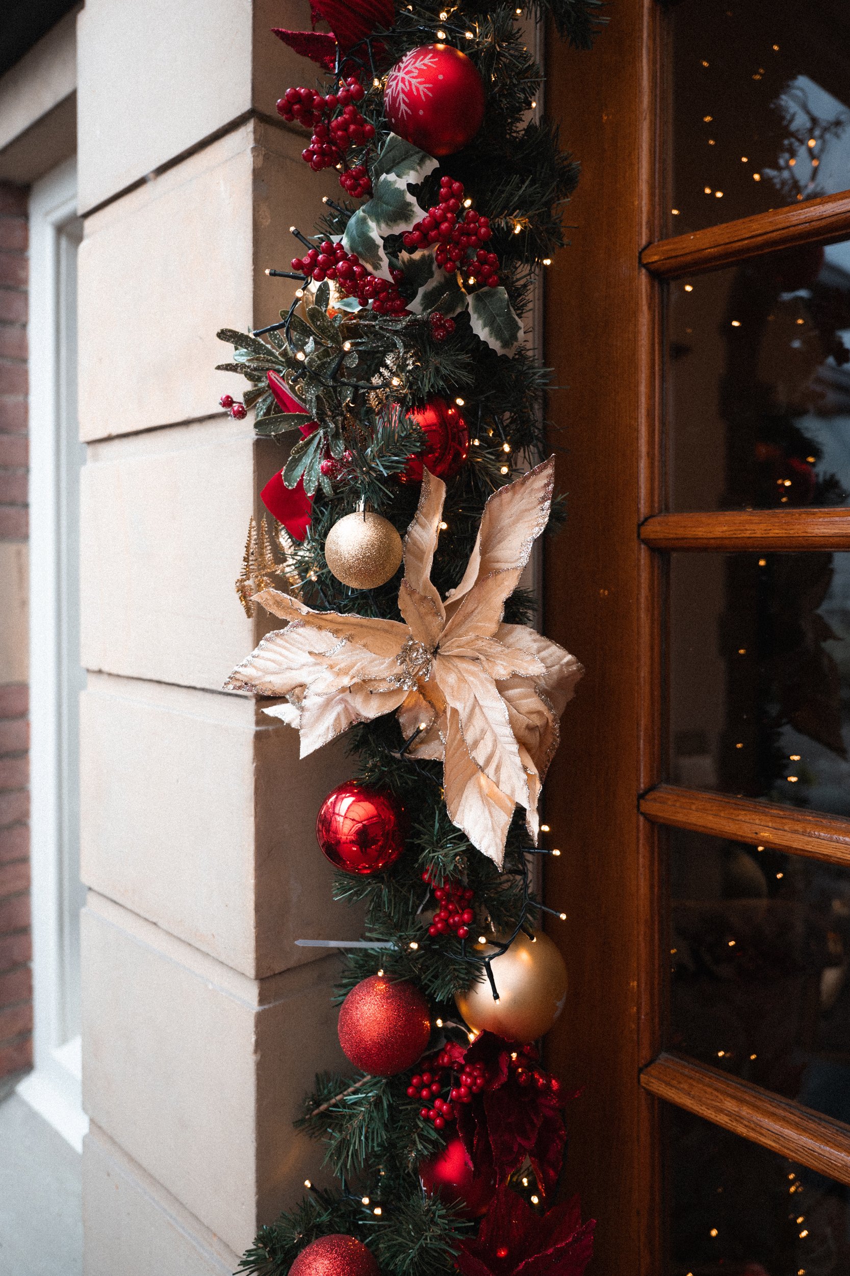 Christmas garland with red and gold ornaments, a large gold poinsettia flower, holly leaves, red berries, and tiny string lights, hanging along a house exterior next to a wooden door.