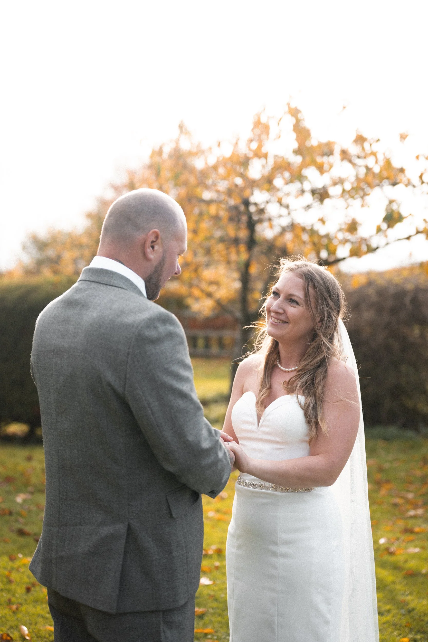A bride and groom exchanging vows outdoors during autumn, with trees in fall colors in the background.