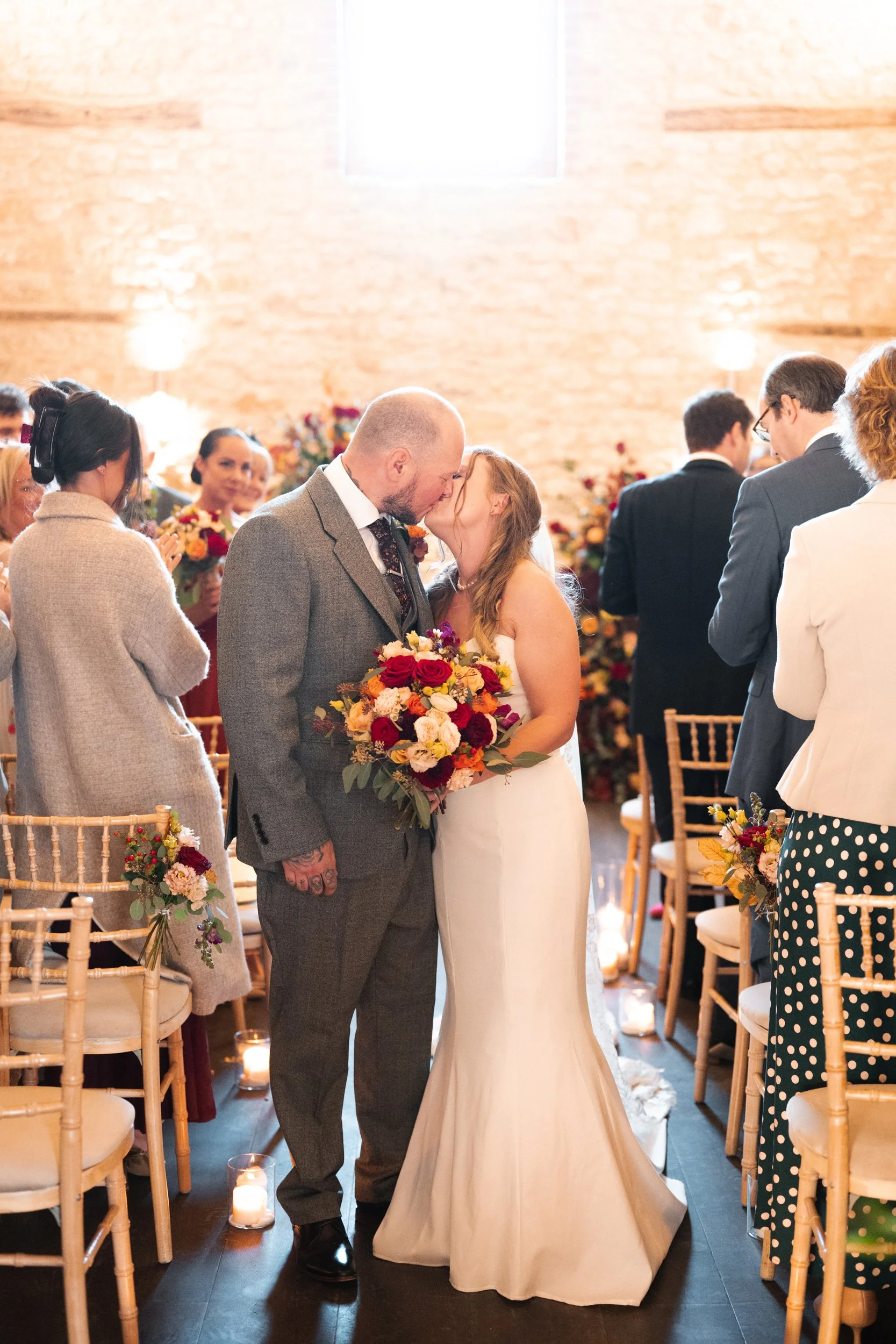 Bride and groom kissing at their wedding ceremony, with guests watching and holding bouquets, in a warmly lit venue with candles and floral decorations.
