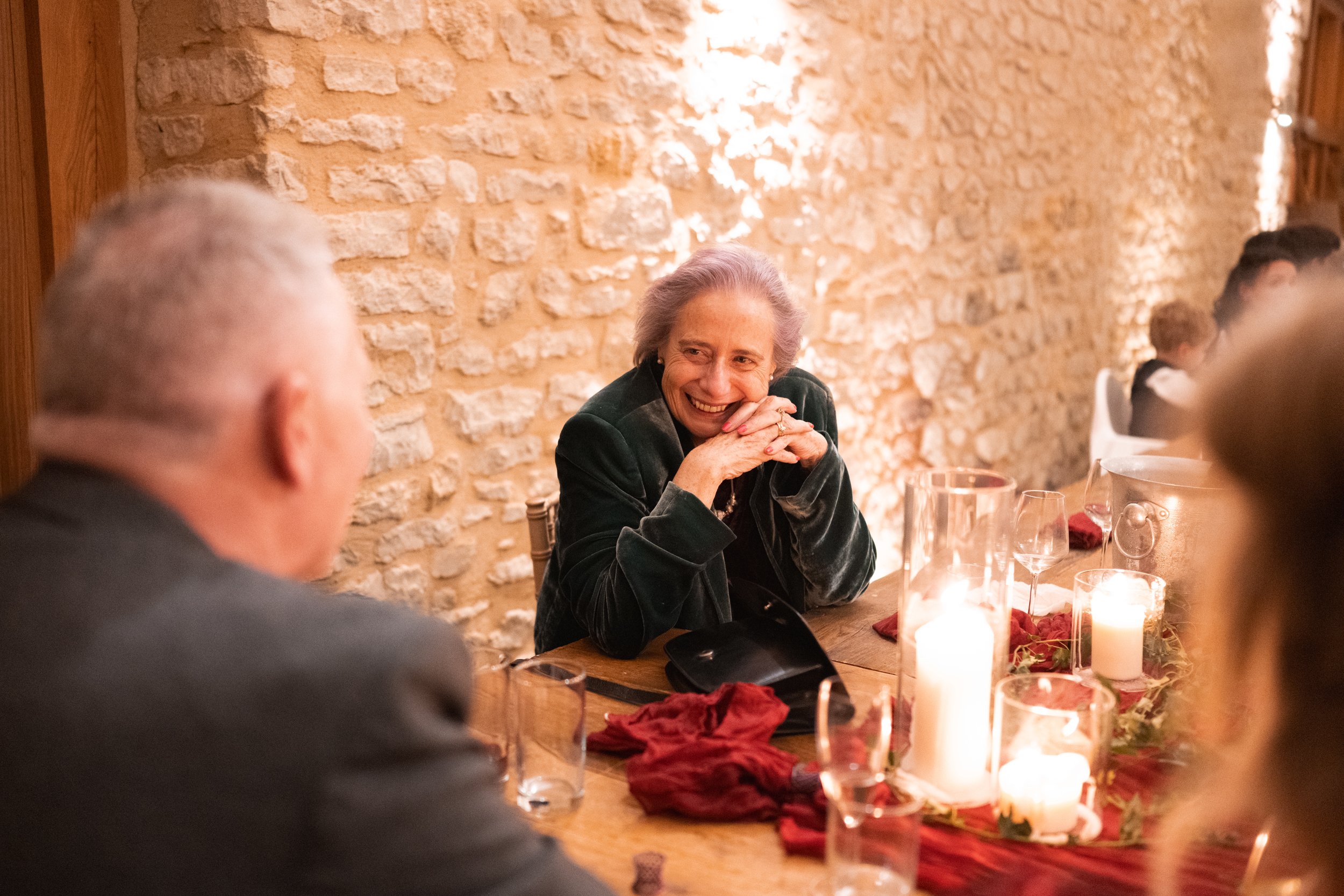 A woman with gray hair smiling at a dining table during a candlelit dinner with a brick wall in the background.