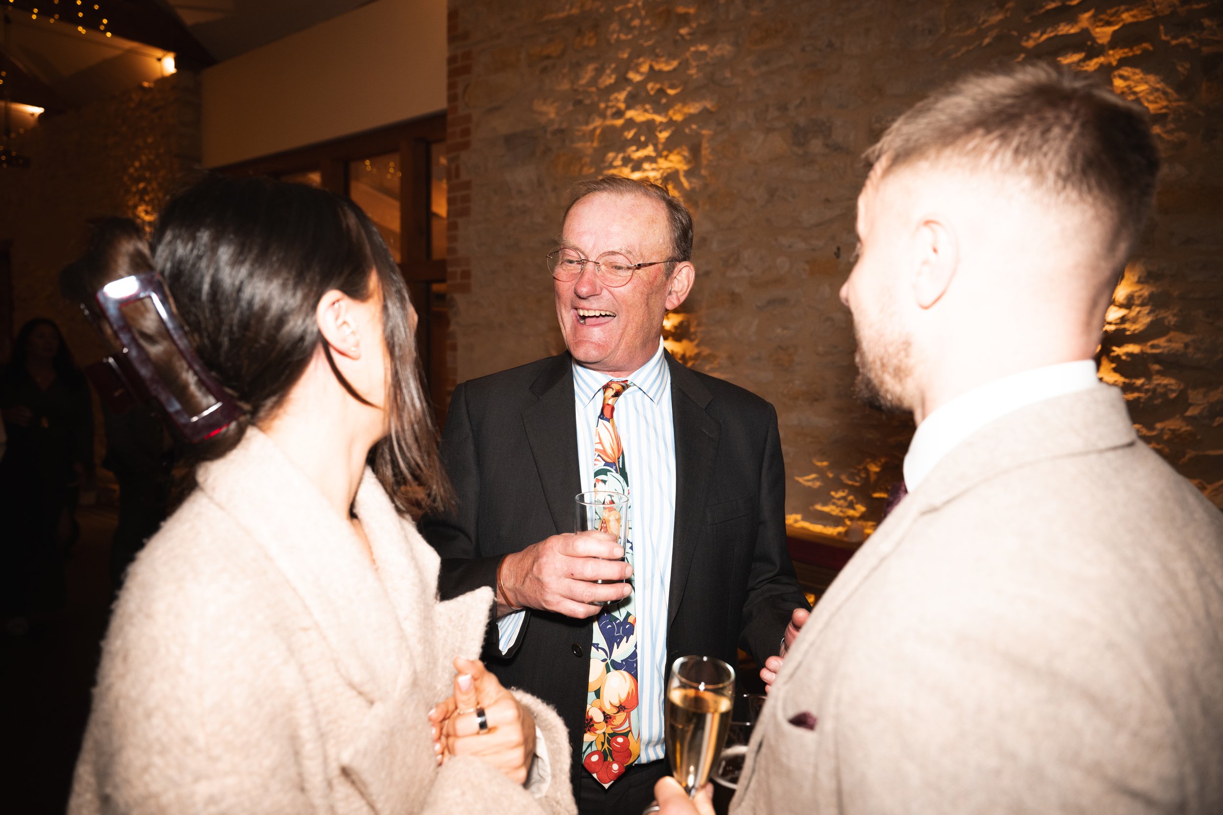Three people at a social gathering, two men and one woman, are engaged in conversation and smiling, with drinks in their hands, in a warmly lit indoor setting.