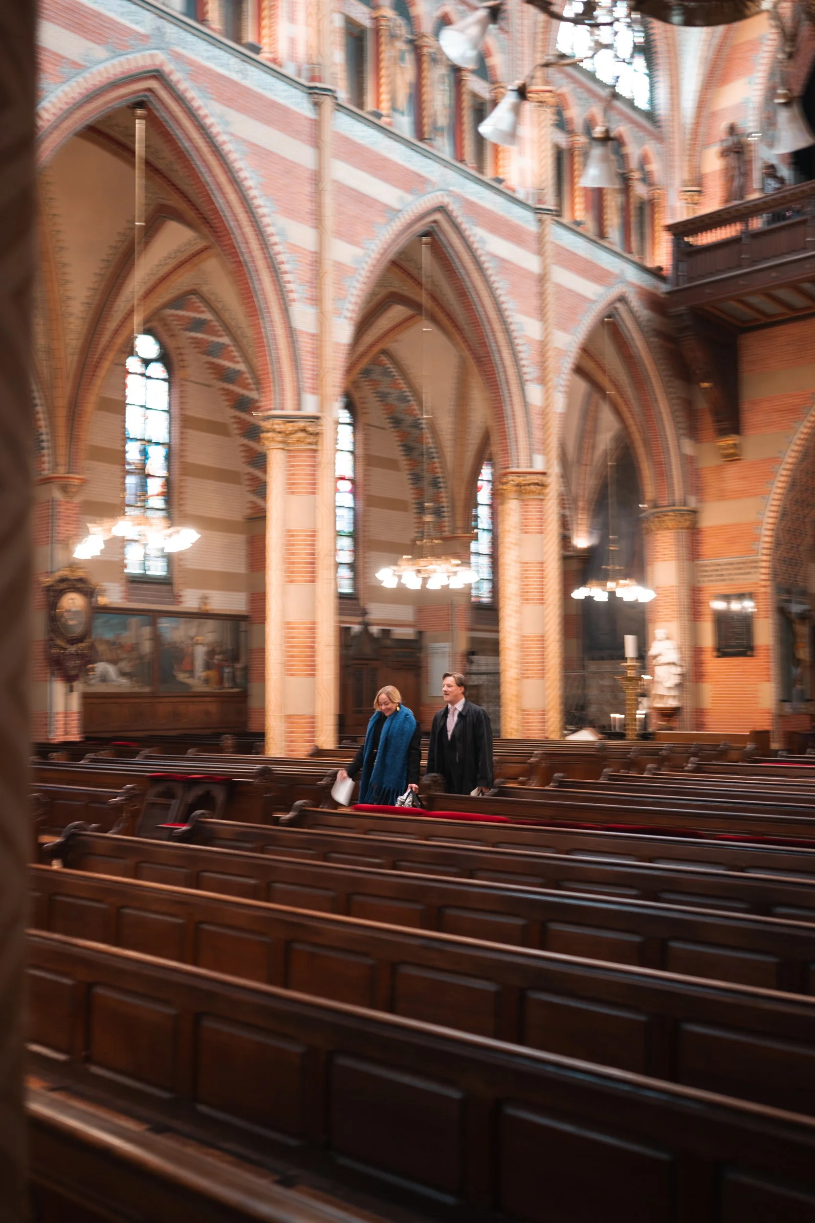 Inside a large, historic church with high, vaulted, brick and stone arches, stained glass windows, and wood pews. Two people are walking down the aisle.