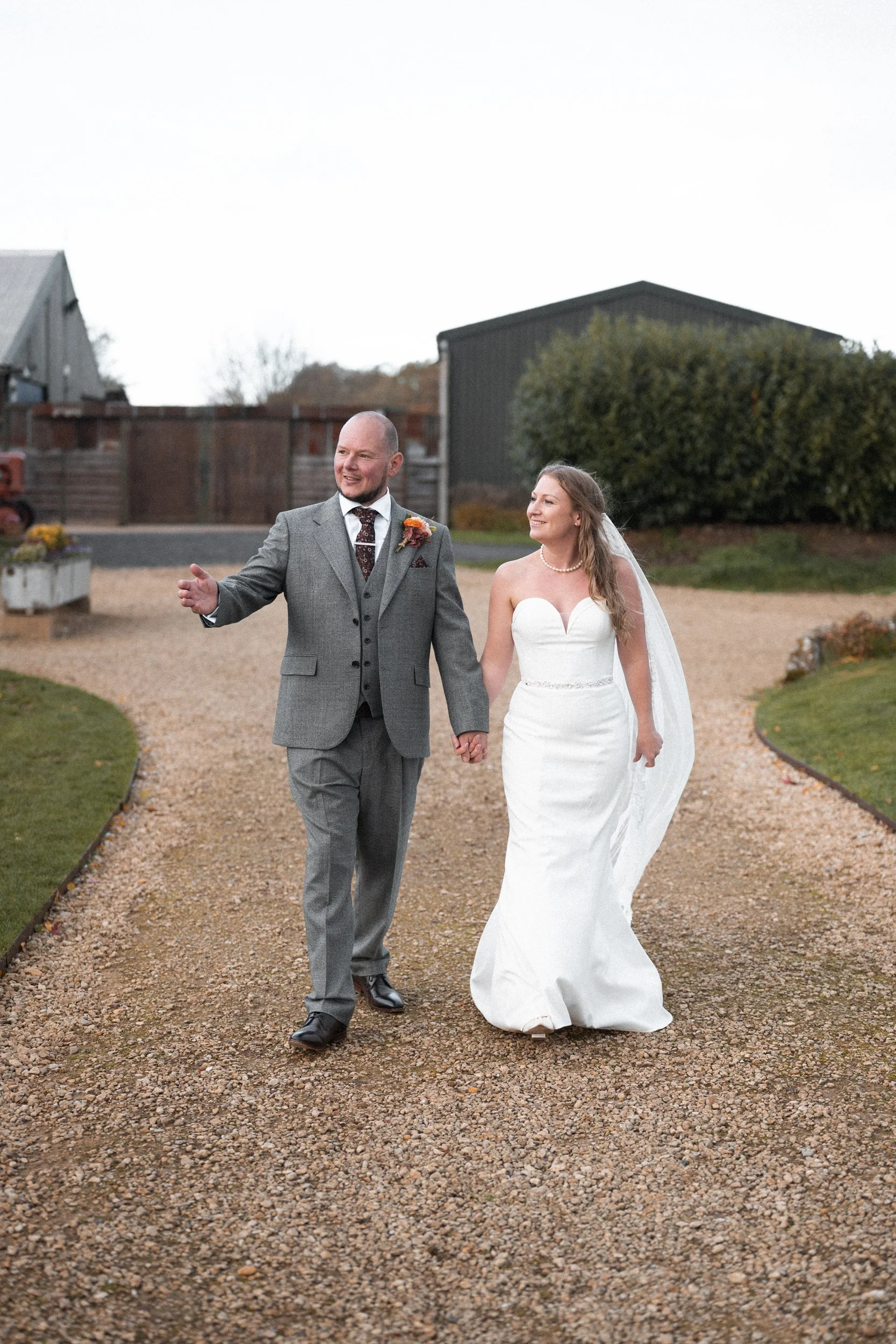 A bride and groom walking hand in hand outside on a gravel path during a wedding celebration, with the groom dressed in a gray suit and the bride in a white strapless wedding gown.