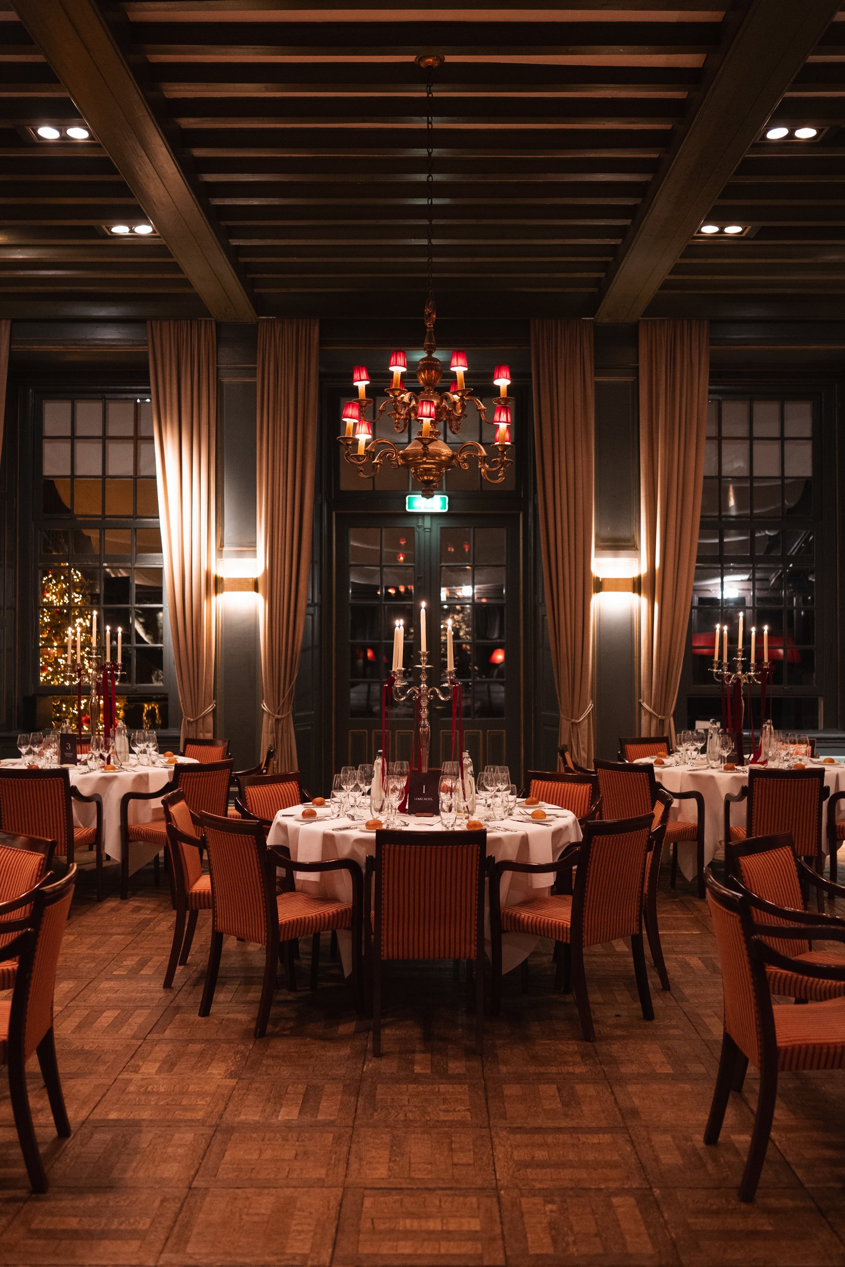 Elegant dining room with round tables covered in white tablecloths, set with wine glasses and silverware, surrounded by upholstered chairs, decorated with tall candlesticks, chandelier with red lampshades, large windows with beige curtains, and holid