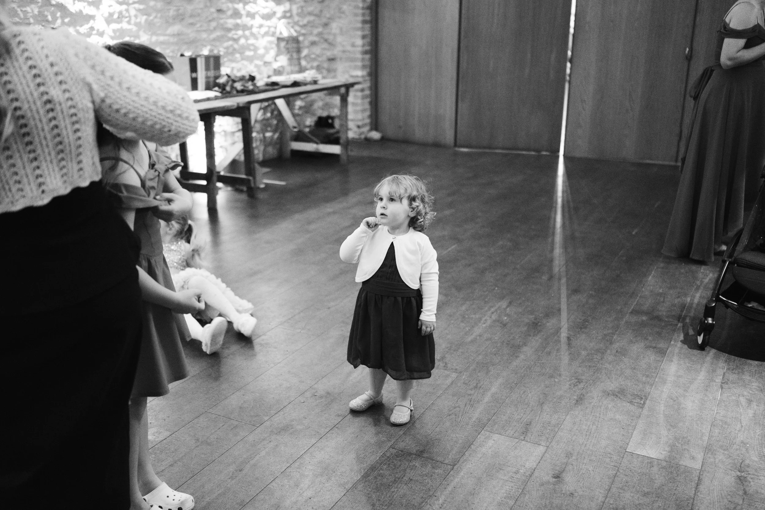A young girl with curly hair wearing a dark dress and a light cardigan stands on a wooden floor in a room, looking slightly upward. Several people are partially visible around her, and a table with items and a doll are seen in the background.