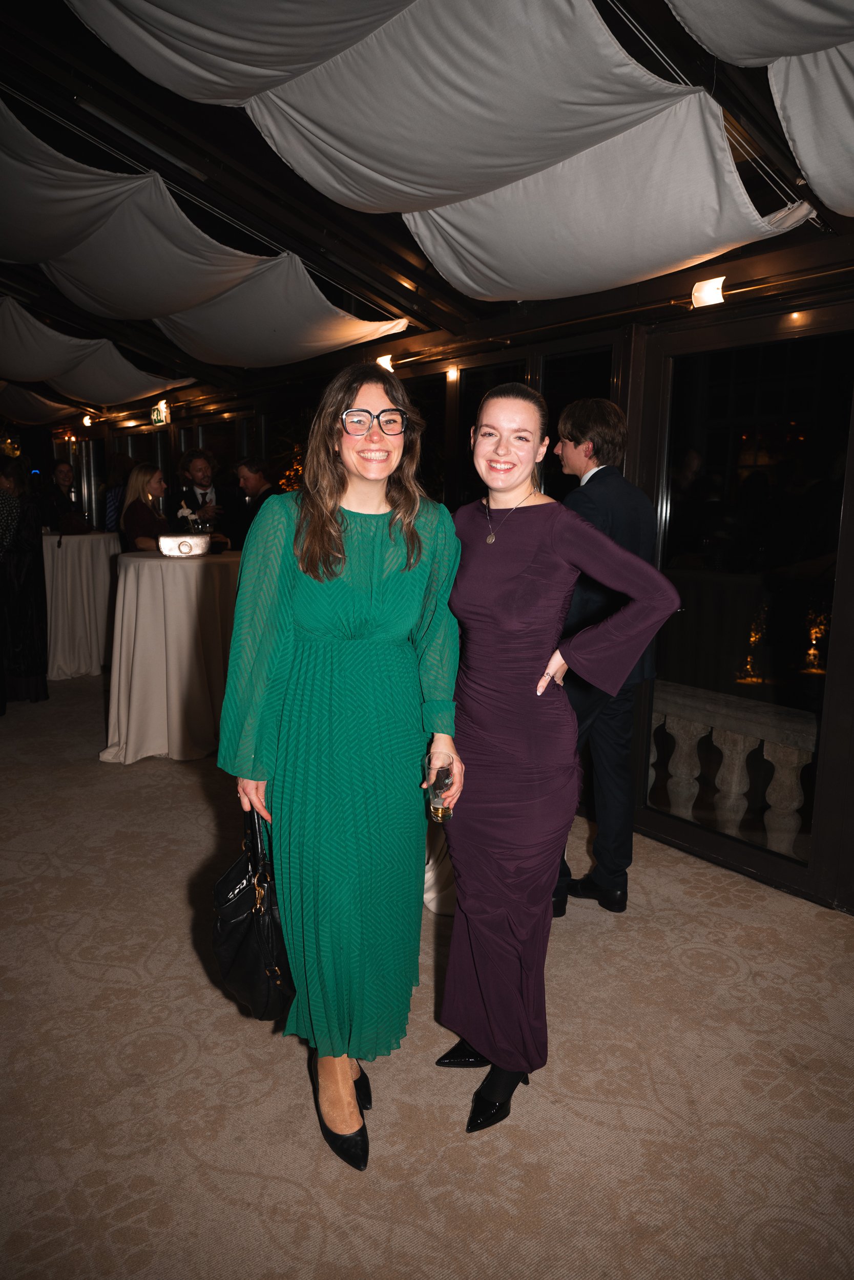 Two women smiling and posing for a photo at a formal event, dressed in elegant dresses, with other guests in the background.