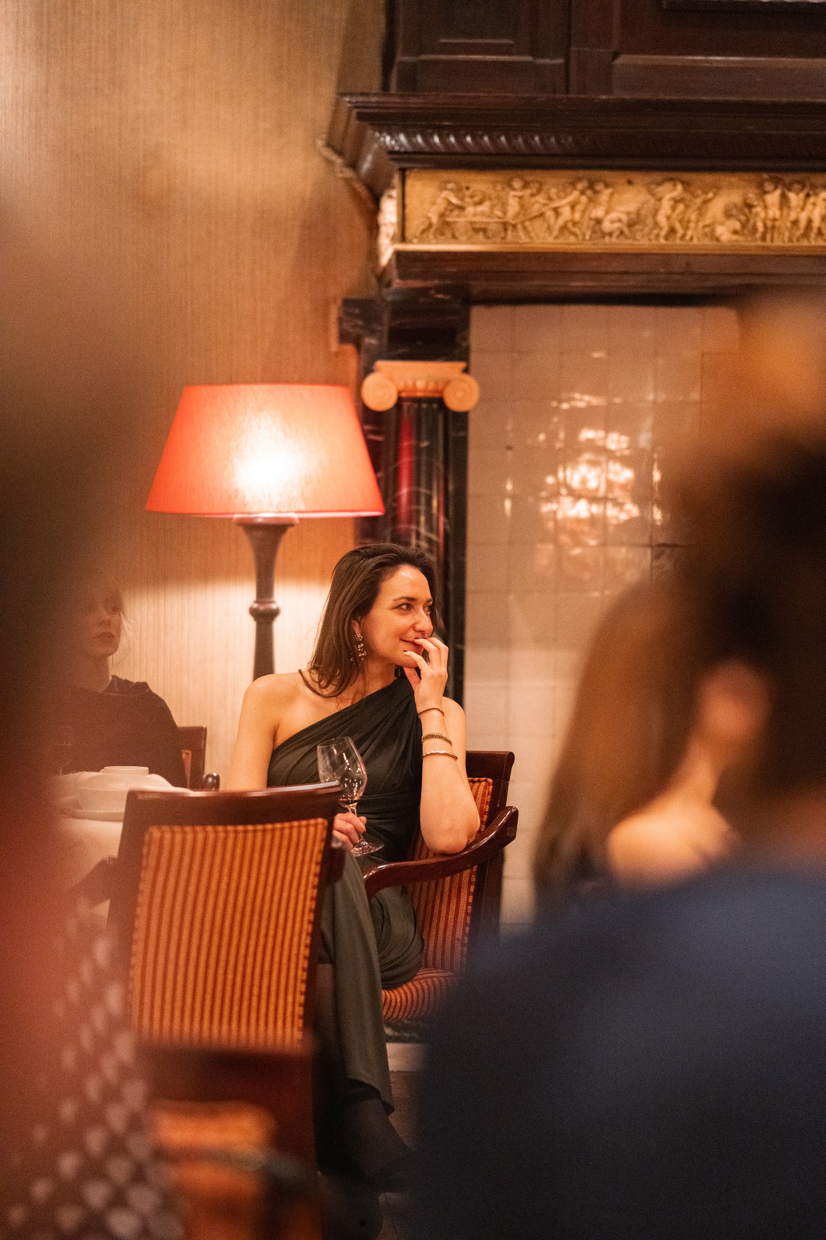 A woman in a black dress is sitting at a restaurant table, holding a wine glass. She is smiling and has her hand near her mouth. The setting is warm and dimly lit, with a table lamp and ornate decorations in the background.