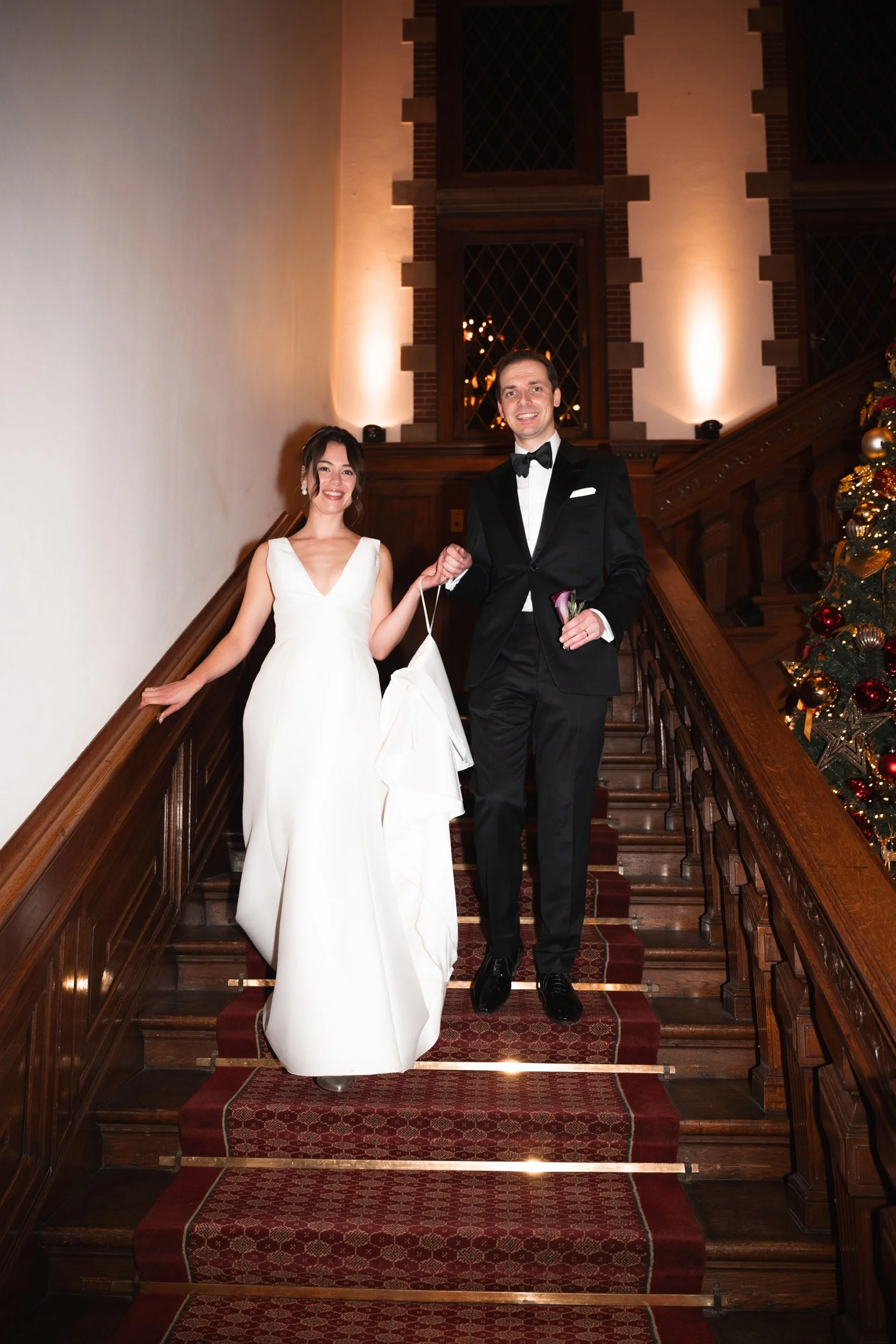 A bride in a white wedding gown and a groom in a black tuxedo walking down a staircase with a wooden railing, holding hands, smiling, and carrying a white jacket. A decorated Christmas tree is visible on the right side of the staircase.
