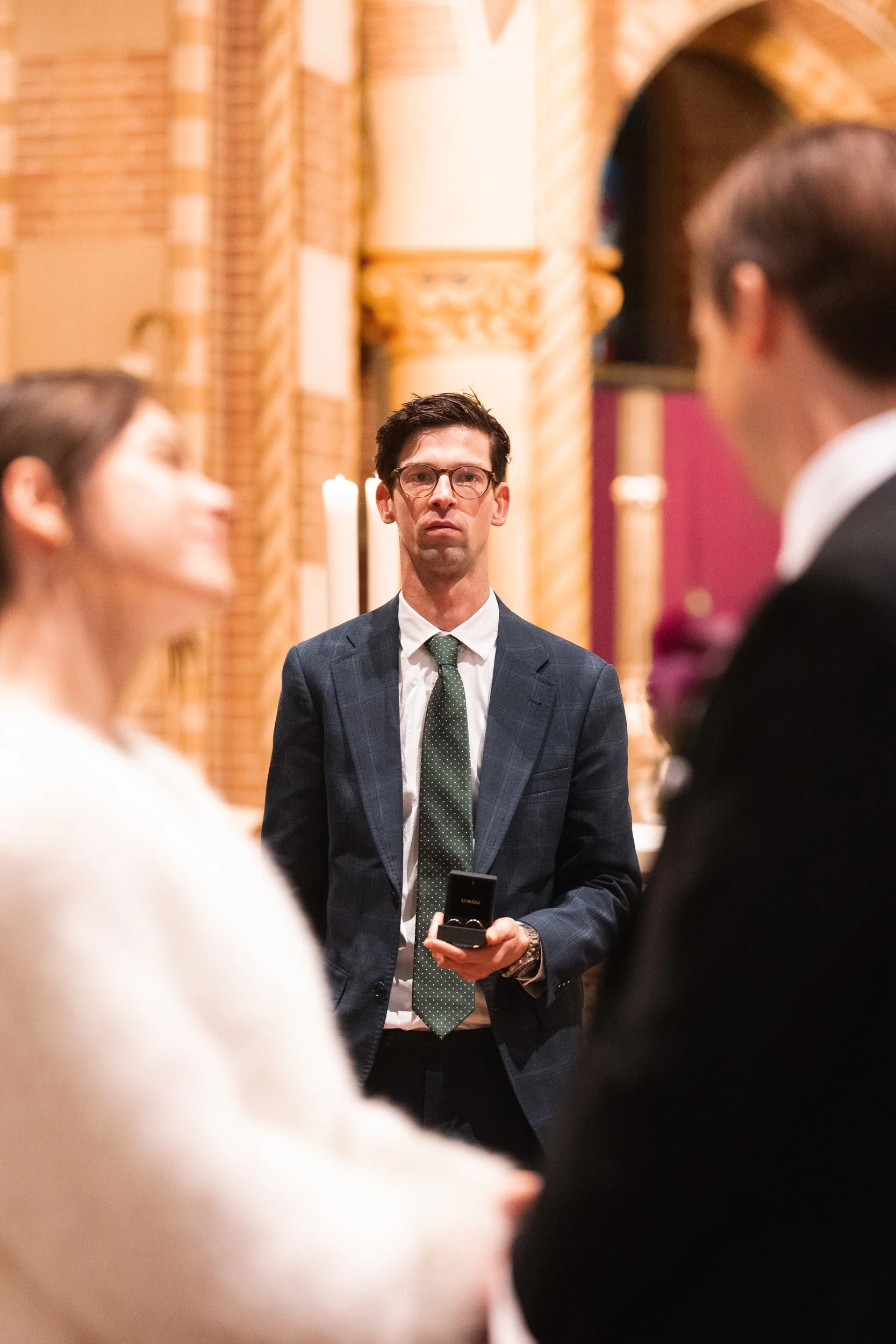 Man in a dark suit, green tie, and glasses standing during a wedding ceremony in a church, holding a small black box.