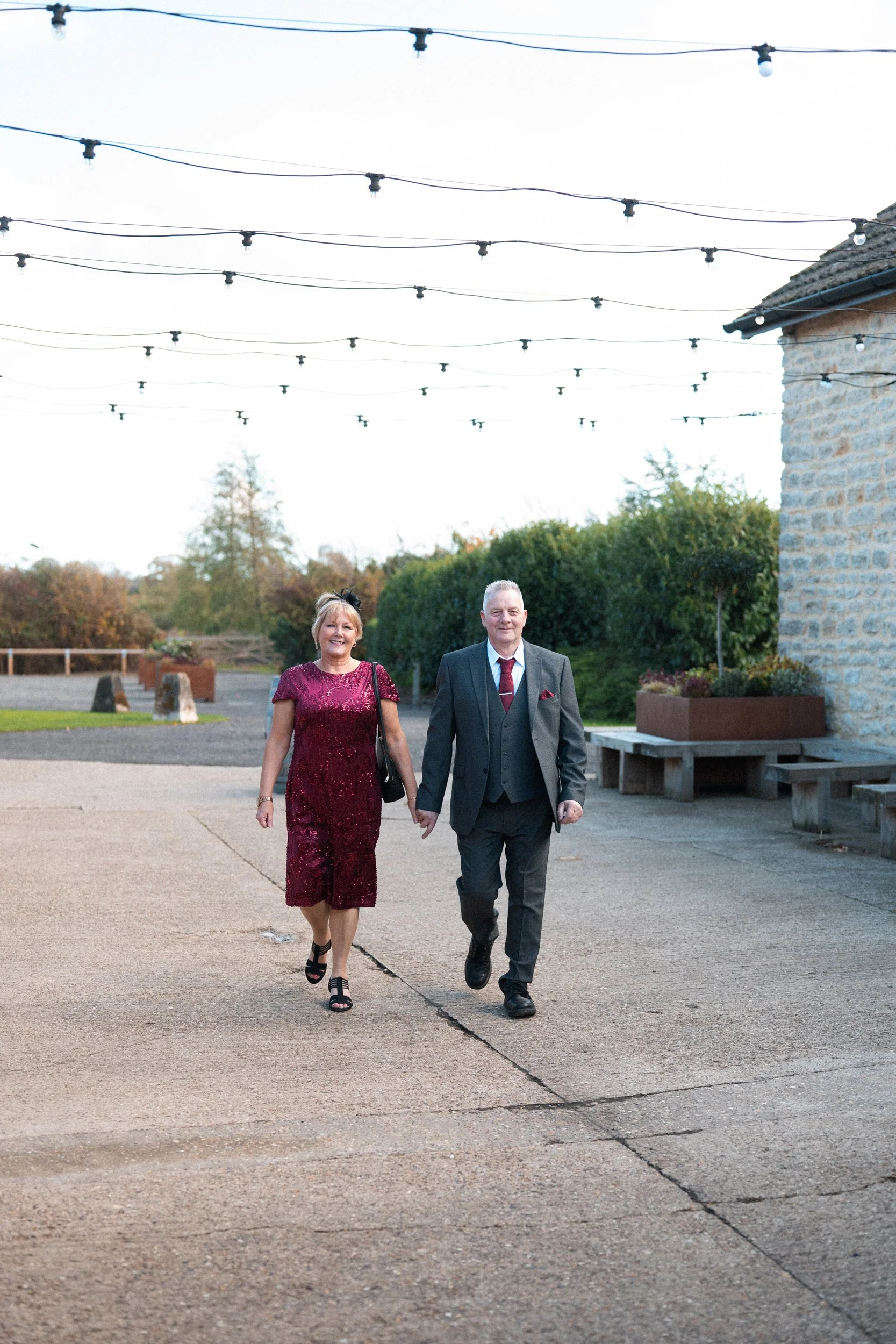 A couple dressed nicely, holding hands and walking outdoors, with string lights overhead and a stone building on the side.