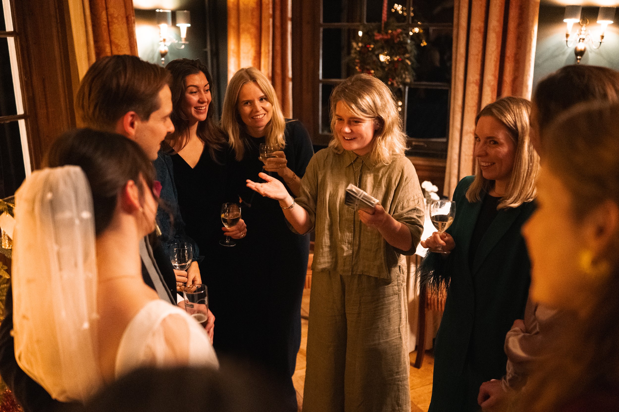 Group of people at a celebration, with a bride in a wedding dress and veil, gathered around a woman who is speaking and holding a stack of papers, in a warmly lit room with curtains and a Christmas tree in the background.