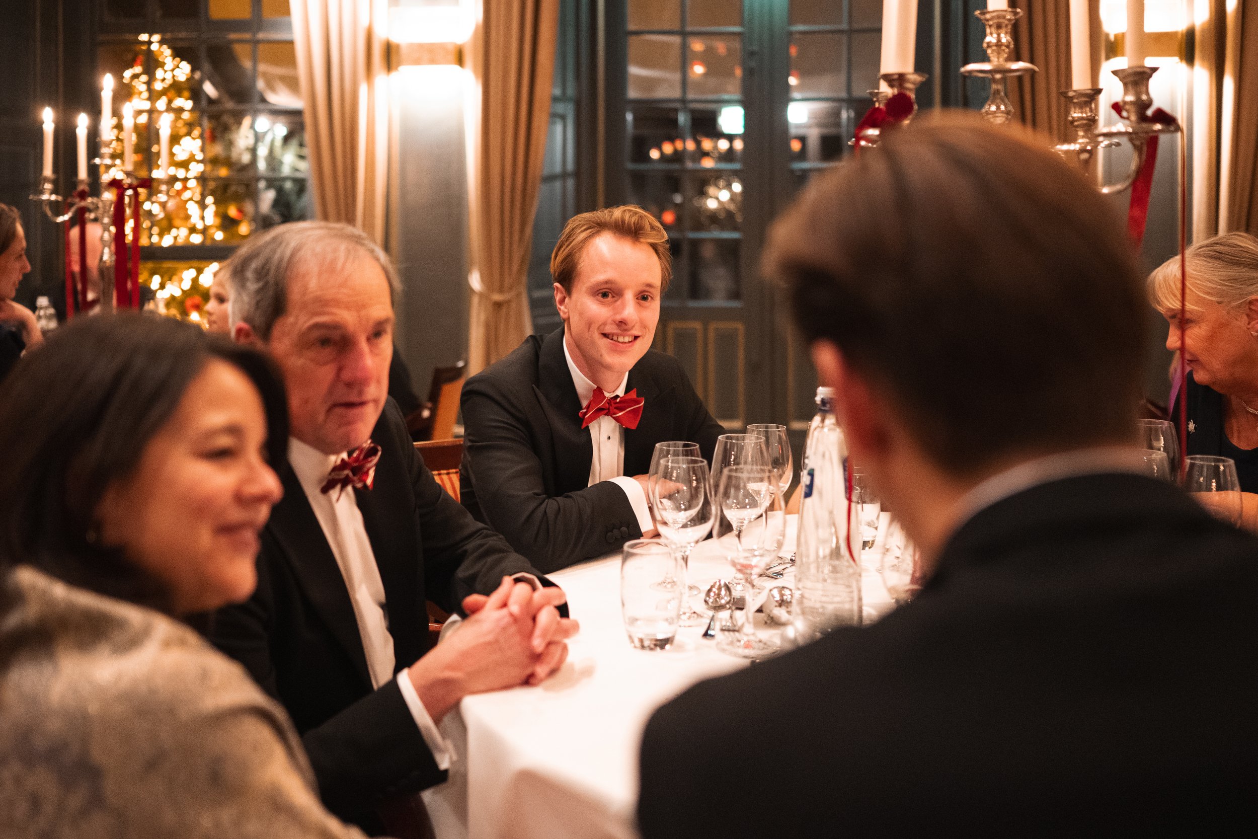 People dressed in formal attire, sitting at a dinner table during a holiday celebration with Christmas decorations, candles, and a Christmas tree in the background.
