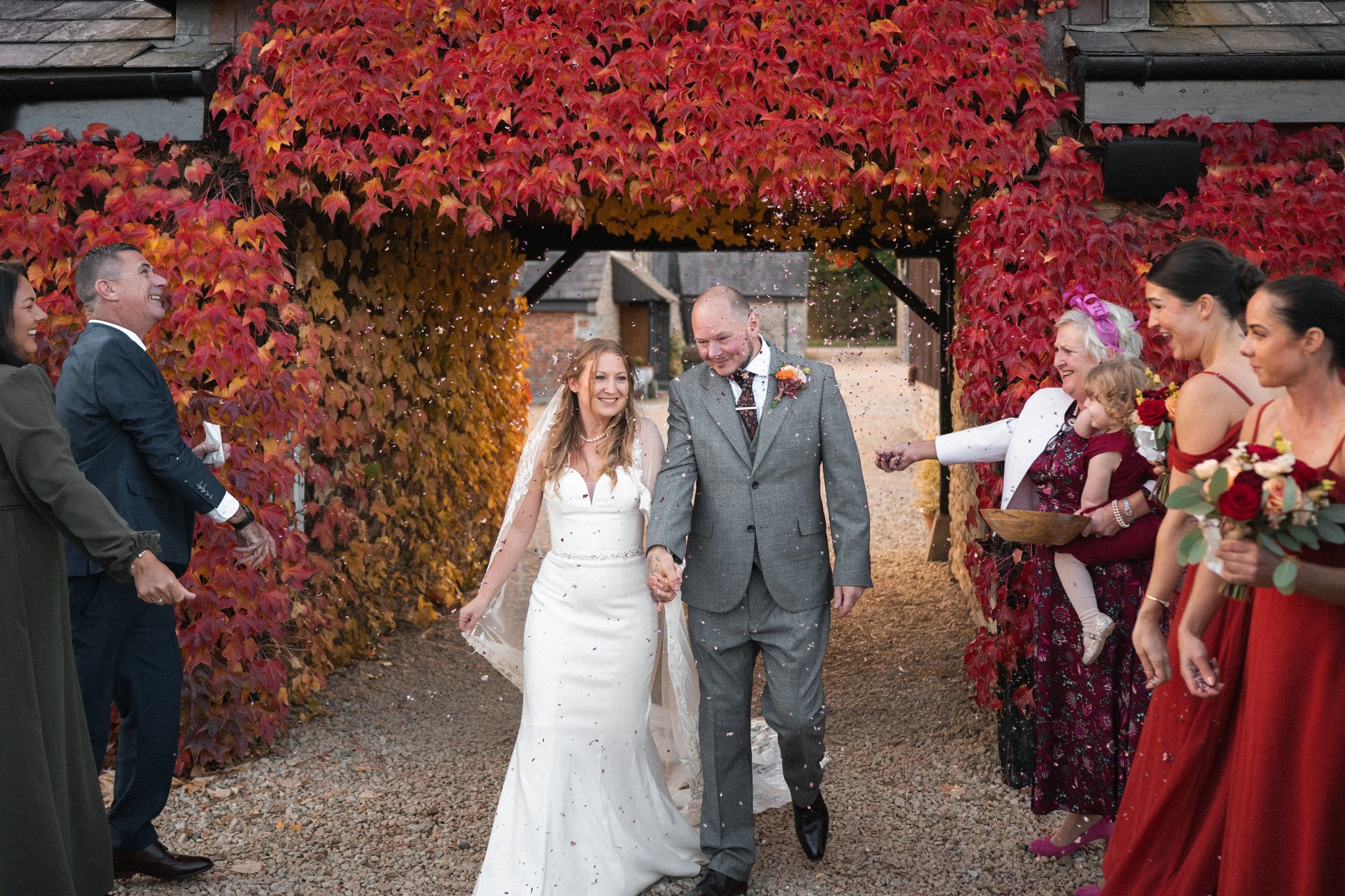 A newlywed couple holding hands walking under an arch covered in red and orange autumn leaves, as friends and family throw confetti in celebration.
